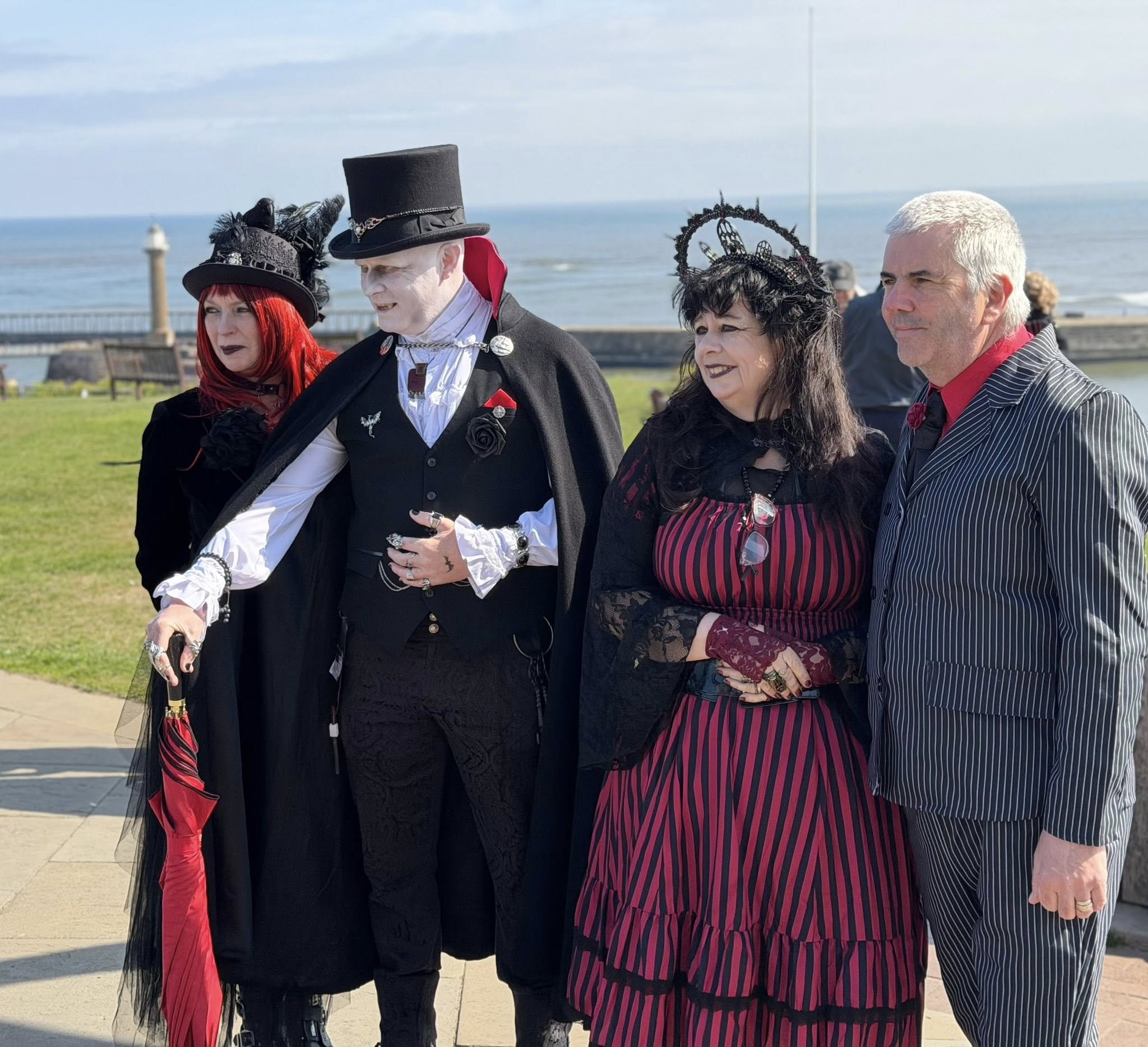 Four people in Victorian and gothic costumes pose beside the ocean, wearing top hats, feathered hats, striped clothing, and dark formal attire.