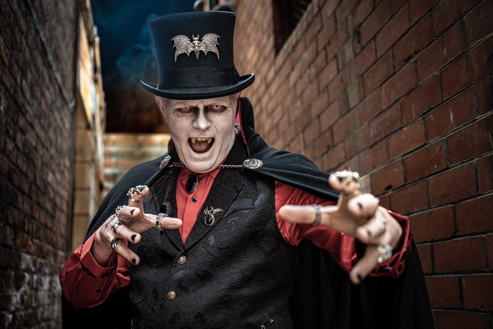 A person dressed as a vampire with pale skin, dark eye makeup, sharp fangs, wearing a tall black hat adorned with a metal bat brooch, a black cape, a red shirt, and layered jewelry, standing in an alleyway made of brick walls.