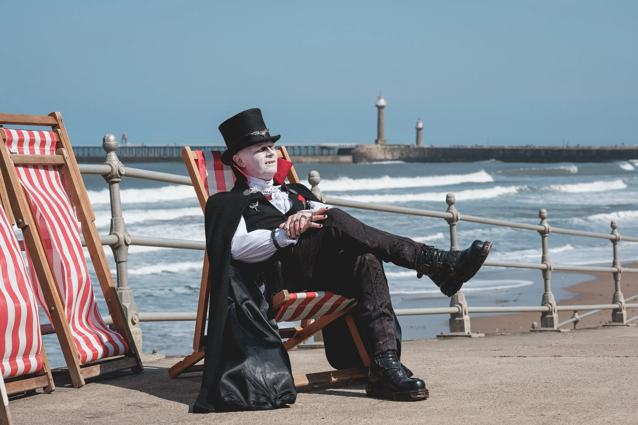 A person dressed as a mime or clown lounging on a striped beach chair at the seaside, wearing a black top hat and cape, with a lighthouse and ocean waves in the background.