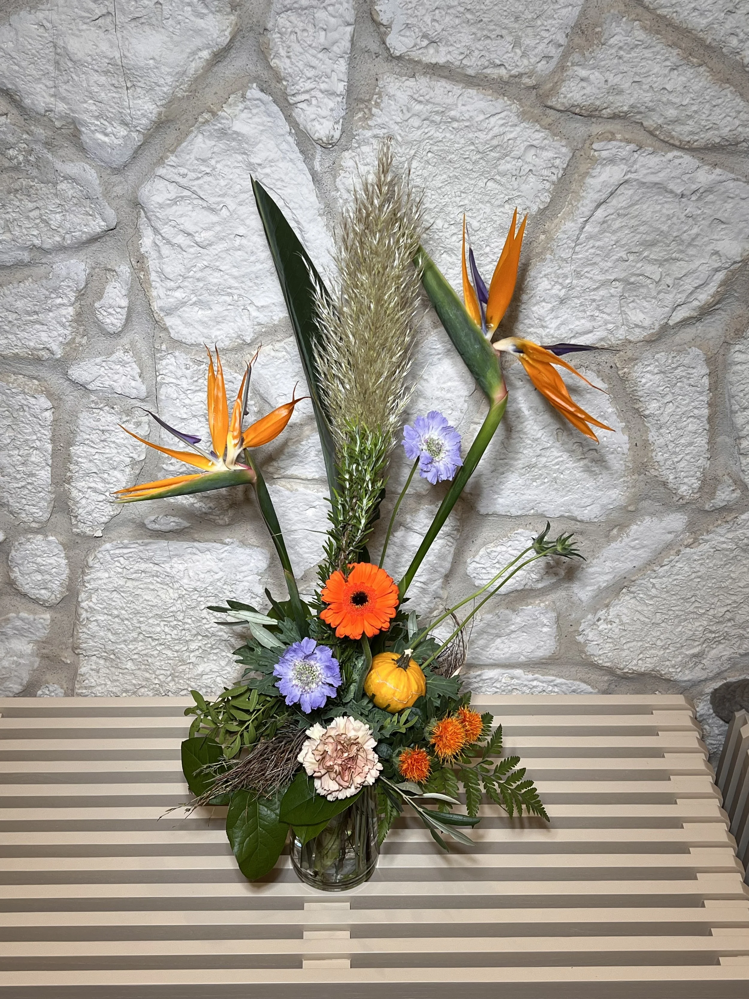 Hohes Herbstarrangement mit Strelitzien, orangefarbener Gerbera, Ziergras und einem kleinen Kürbis.