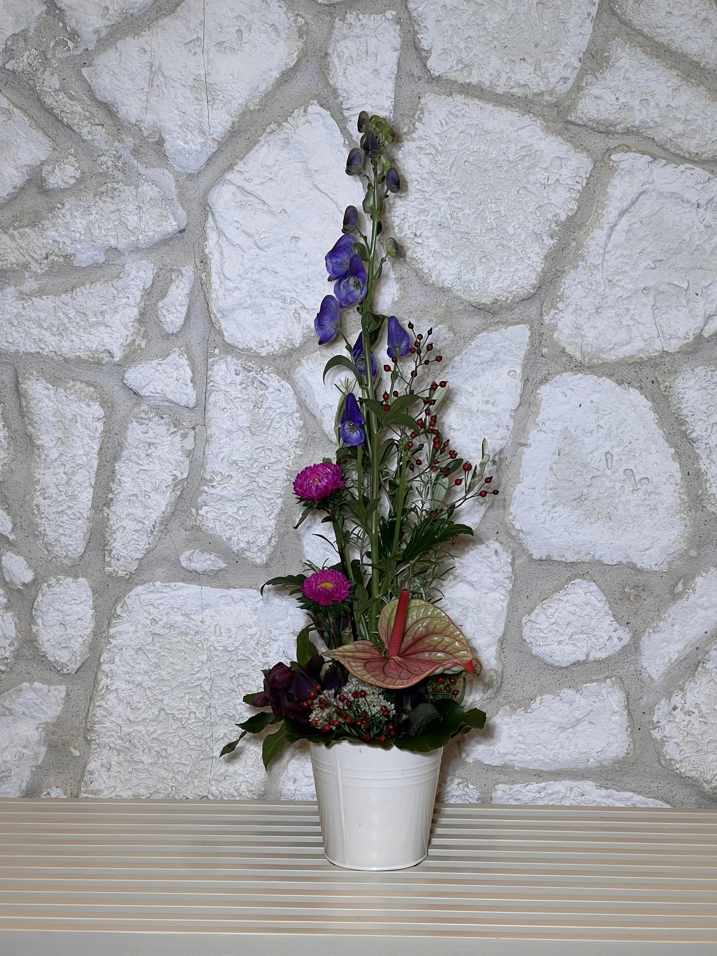 Schlankes Gesteck mit violetten und pinken Blüten, einer Anthurie und roten Beeren in einem weissen Gefäss.