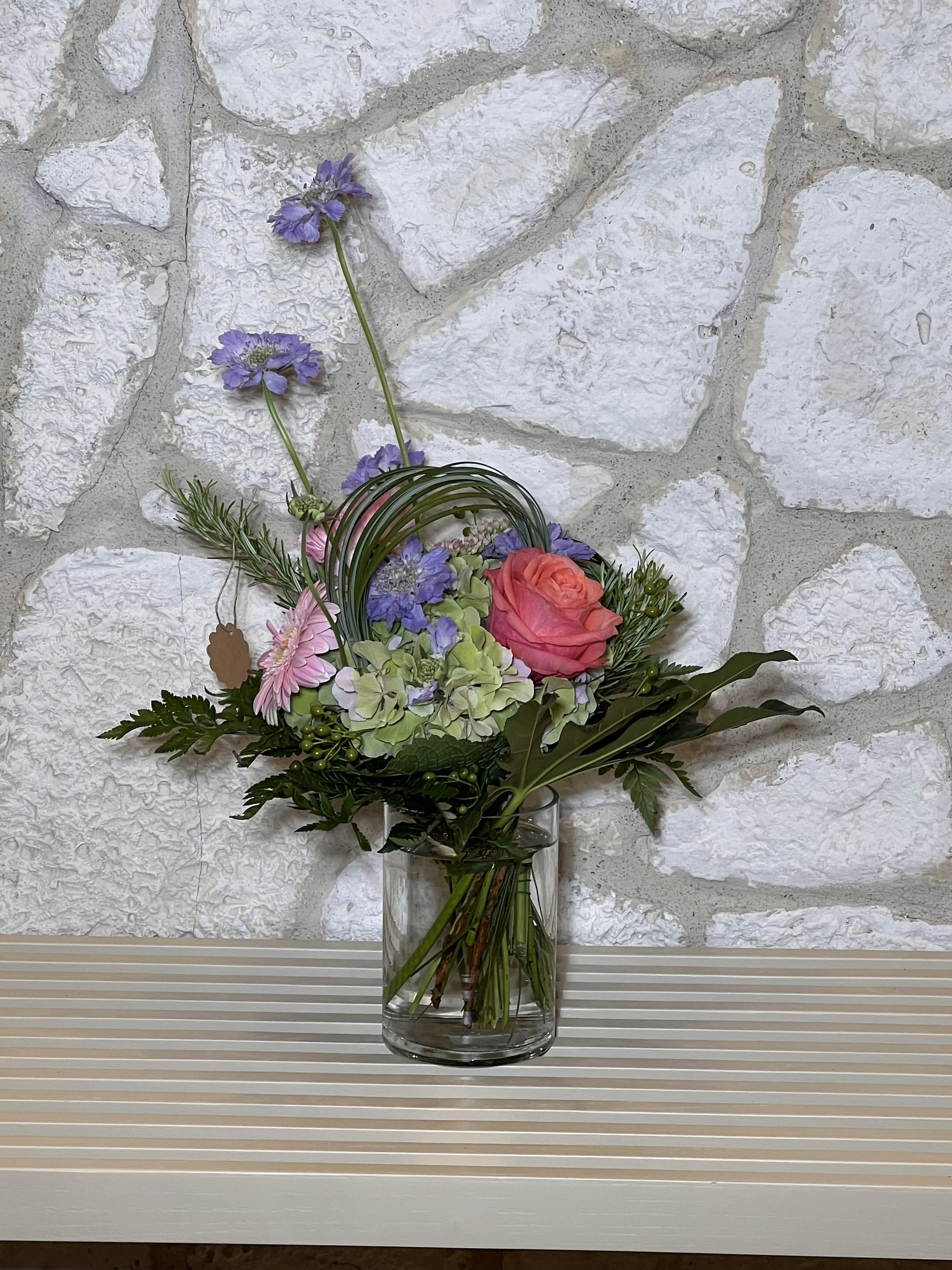 Blumenarrangement mit einer korallfarbenen Rose, grüner Hortensie, violetten Blüten und gebogenen Gräsern in einer Glasvase.