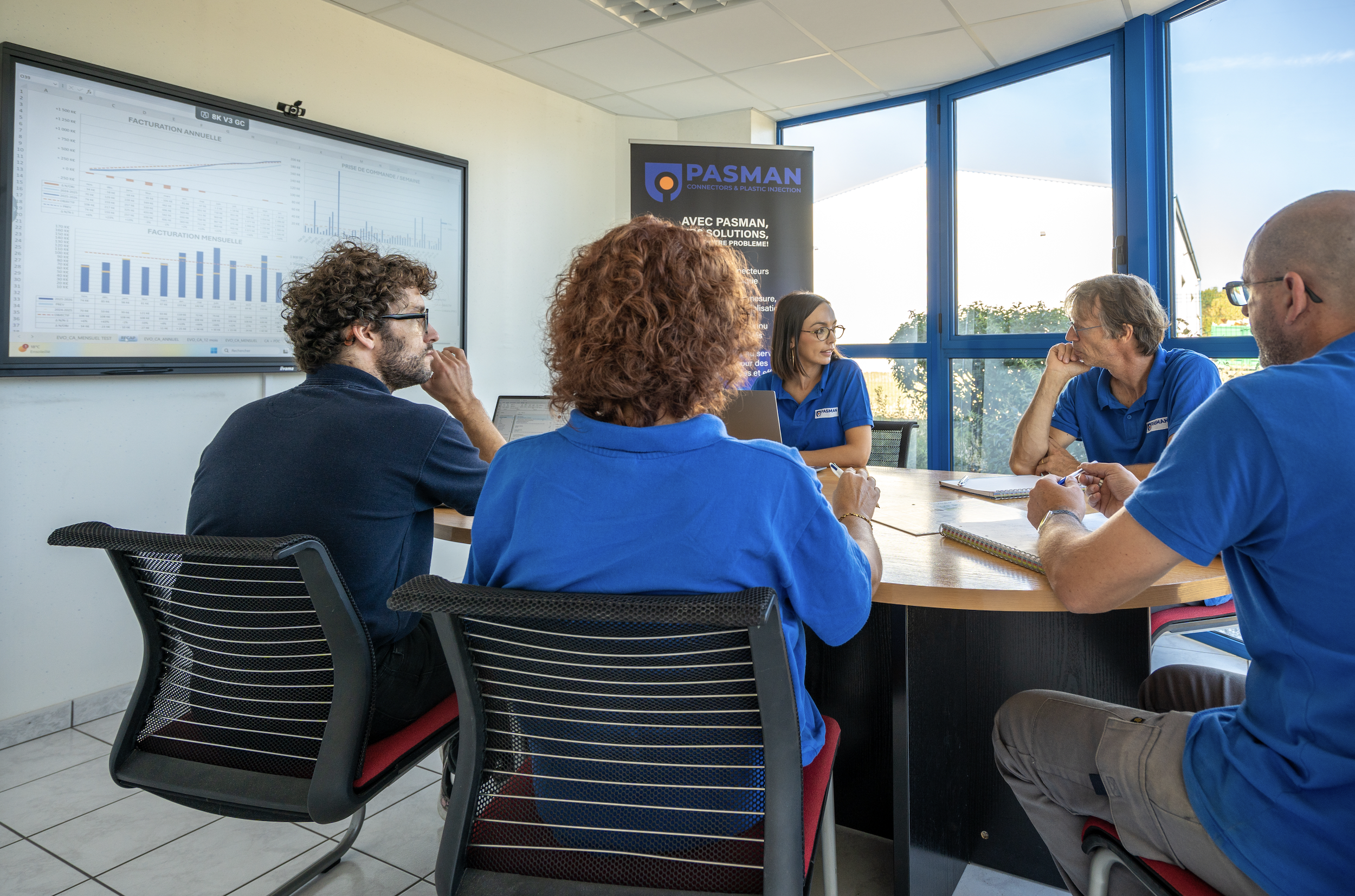Une réunion dans une salle de conférence avec quatre personnes portant des chemises bleues, assises autour d'une table. Un écran affiche des graphiques et des tableaux. Un panneau Publicitaire PASMAN est visible à l'arrière.