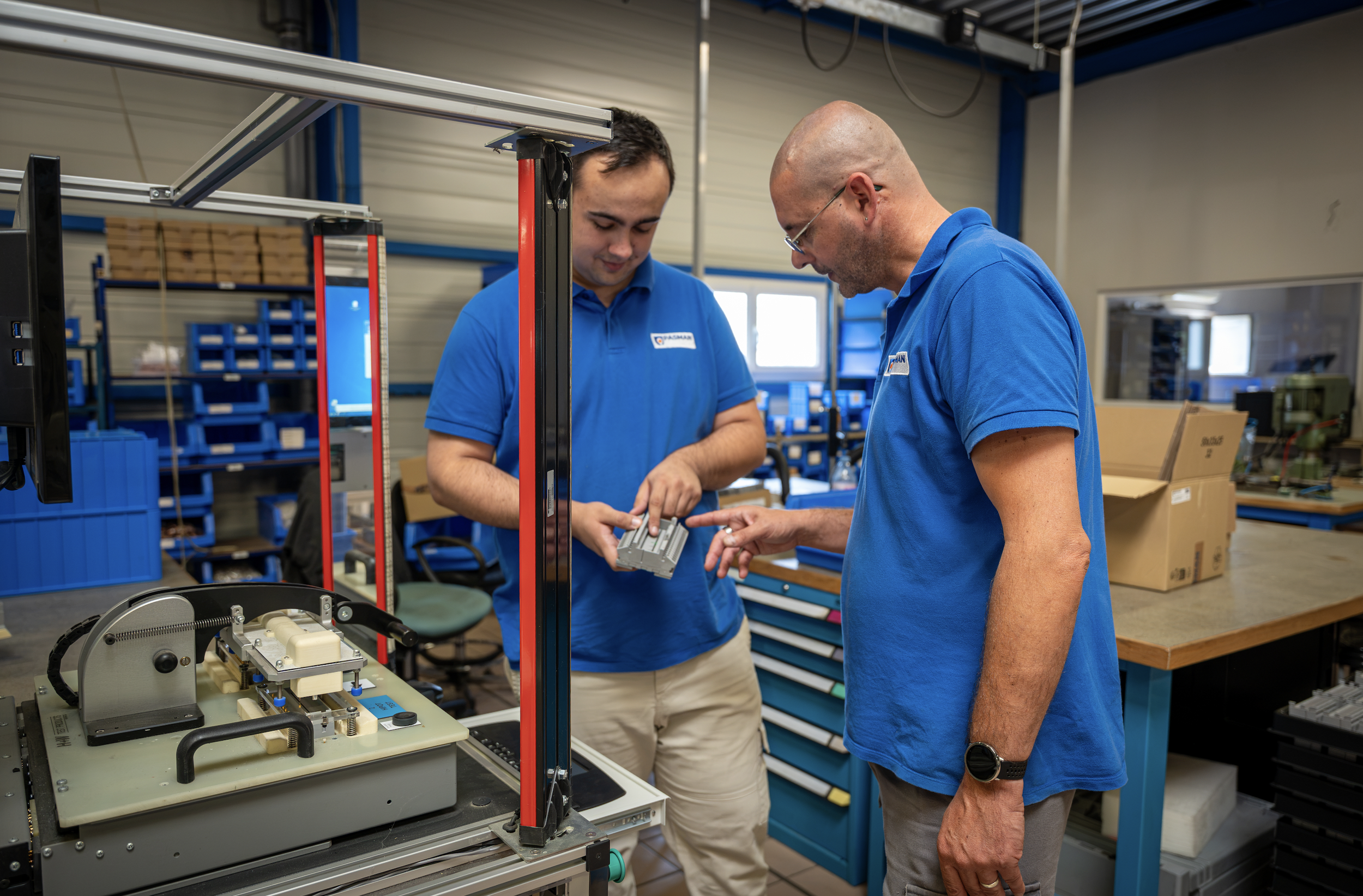 Deux hommes portant des polos bleus travaillent ensemble avec un équipement numérique dans un atelier ou un laboratoire.