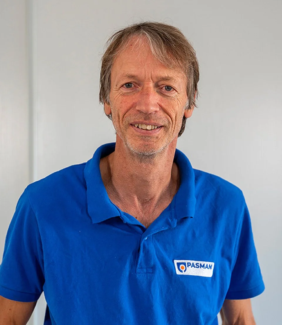 Homme souriant portant un polo bleu avec le logo PASMAN, debout devant un mur blanc.