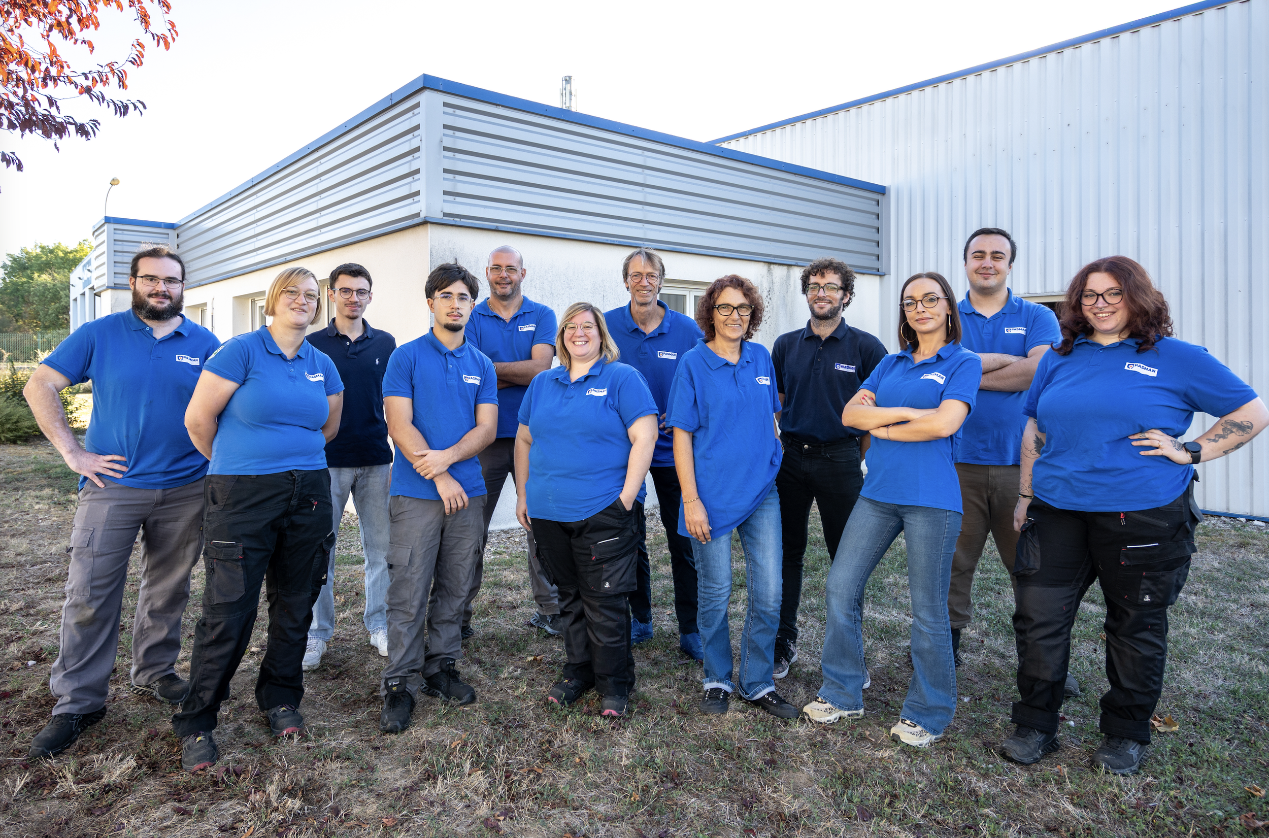 Groupe de dix personnes posant devant un bâtiment industriel, portant toutes des polos bleus ou noirs avec un logo, souriantes et décontractées.