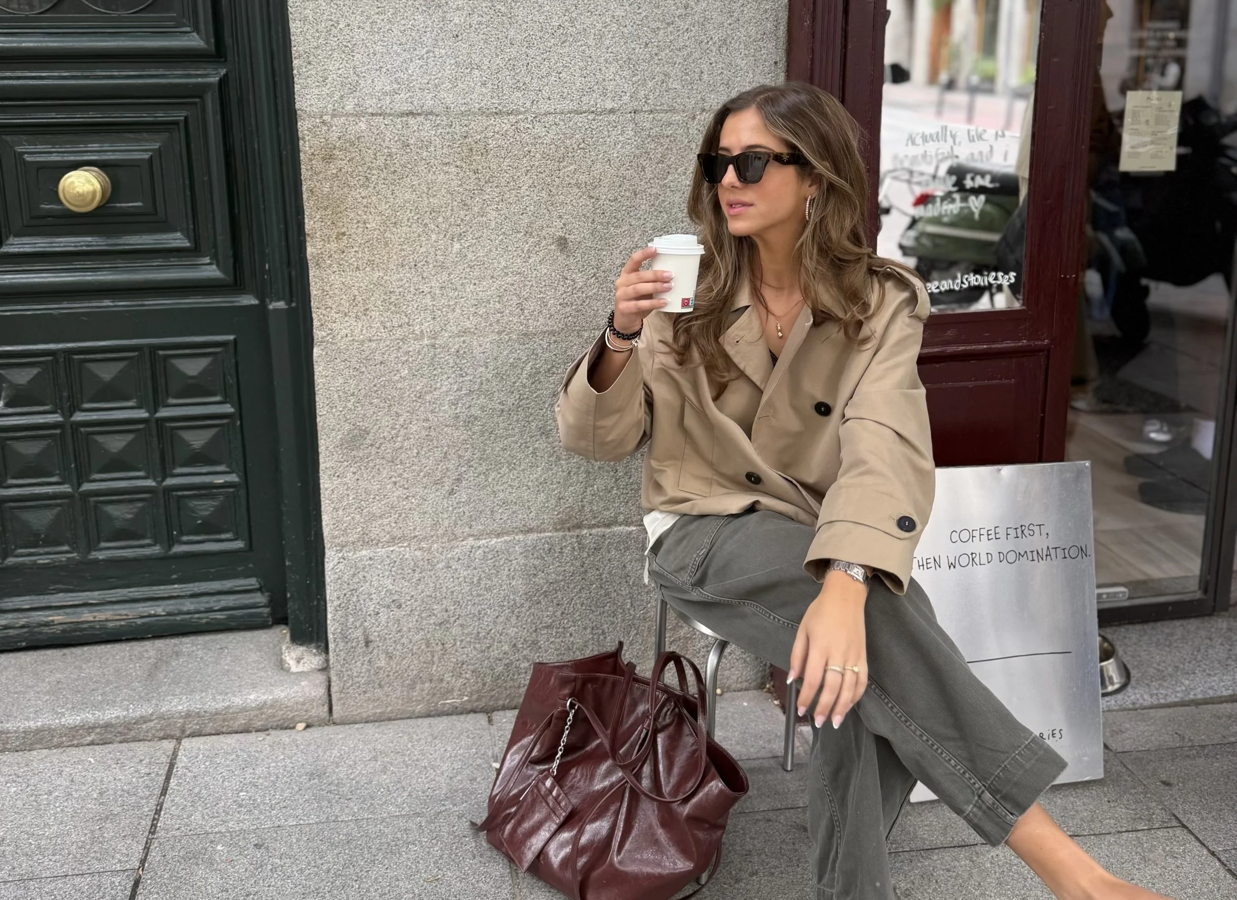 A woman sitting outside a coffee shop with a coffee cup in her hand, wearing sunglasses and a beige trench coat, with a large maroon handbag on the ground beside her. She is in front of a window displaying handwritten signs.