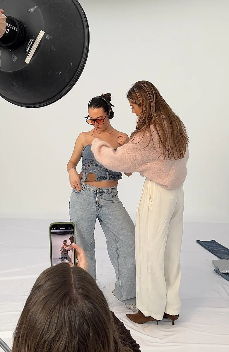 Two women pose for a photoshoot in a studio with white background. One woman wears oversized sunglasses, a denim crop top, and loose jeans. The other woman wears a pink sweater and white pants. A person in the foreground captures the scene on a smartphone. A large studio light is visible in the upper left corner.