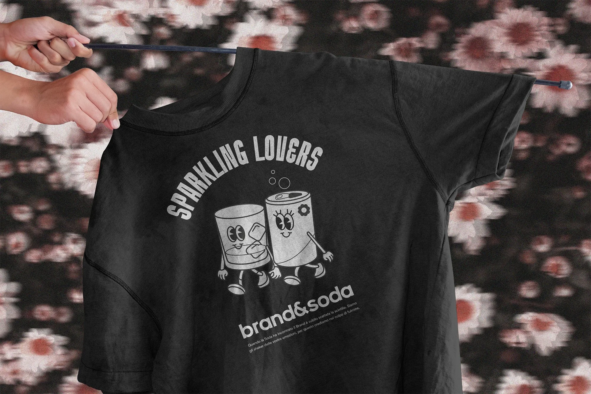 Una maglietta nera con il logo di un'etichetta di soda con due bicchieri che si tengono per mano. La persona che tiene la maglietta la espone contro uno sfondo sfocato.