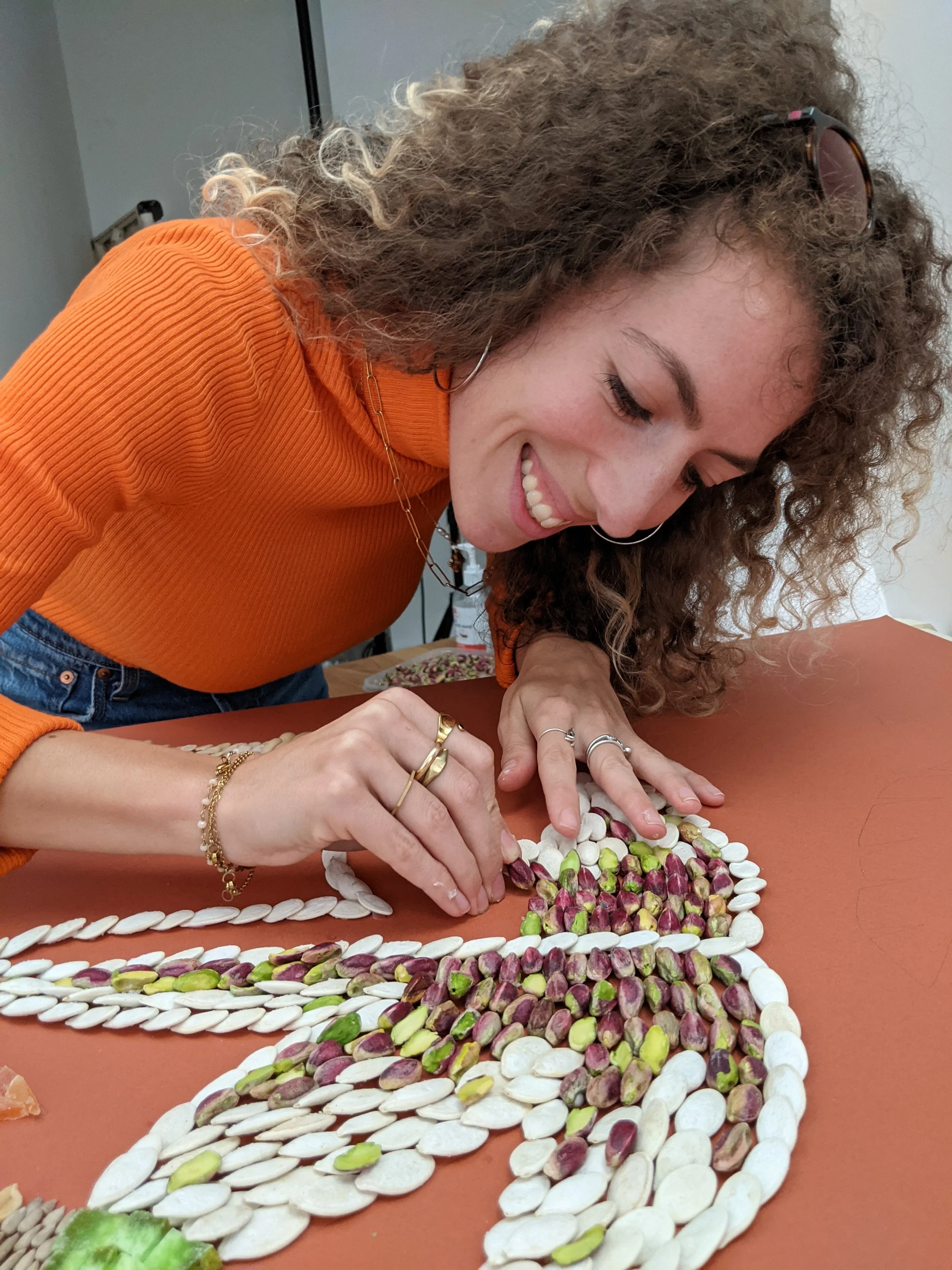 Una donna con capelli ricci e occhiali da sole appoggiati sulla testa, indossa una maglietta arancione e braccialetti dorati, mentre crea un disegno con pistacchi e mandorle su un tavolo di colore marrone.