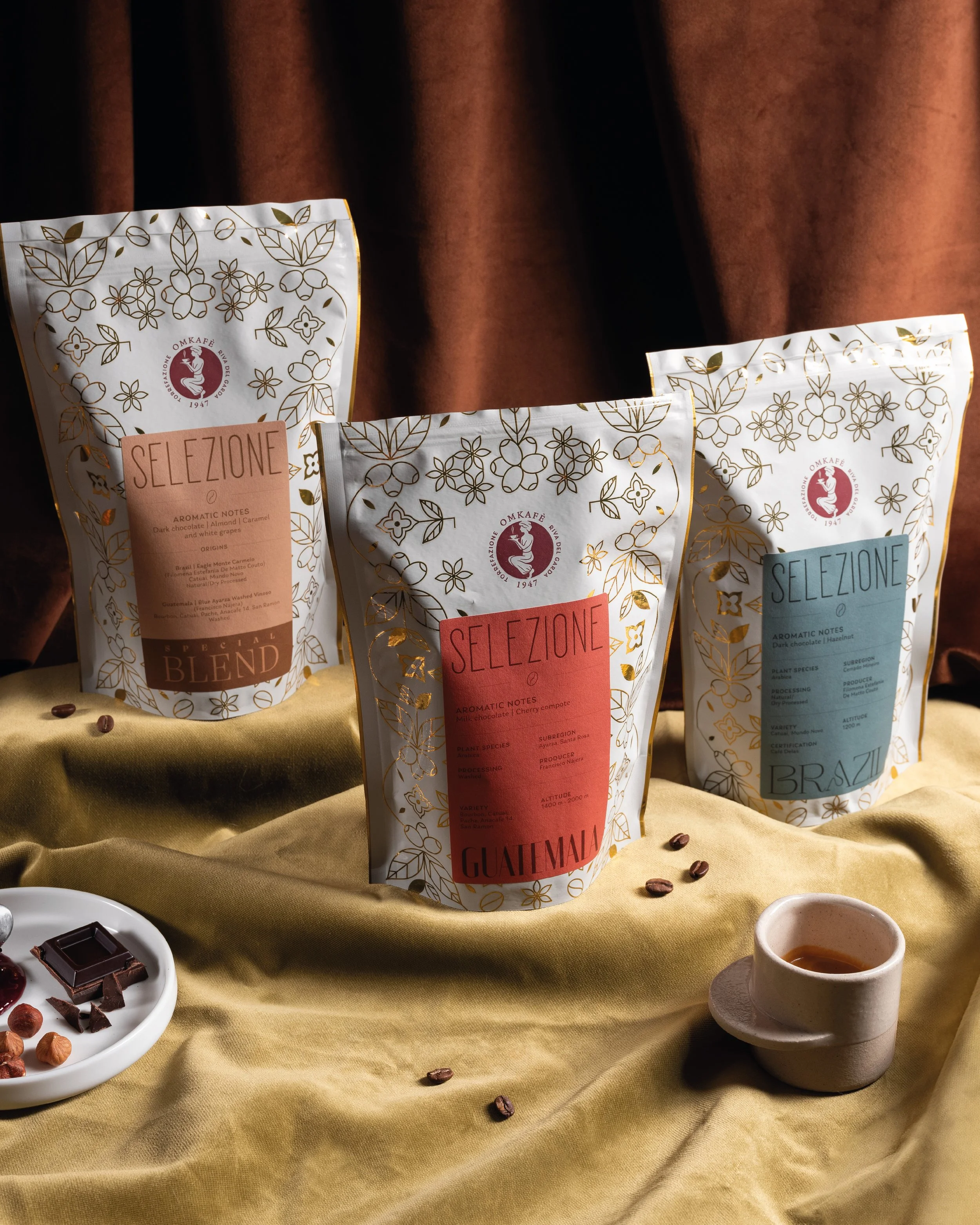Confezioni di caffè in vari colori con etichette di selezione, cioccolato fondente, tazza di caffè, e cioccolatini su tessuto dorato.