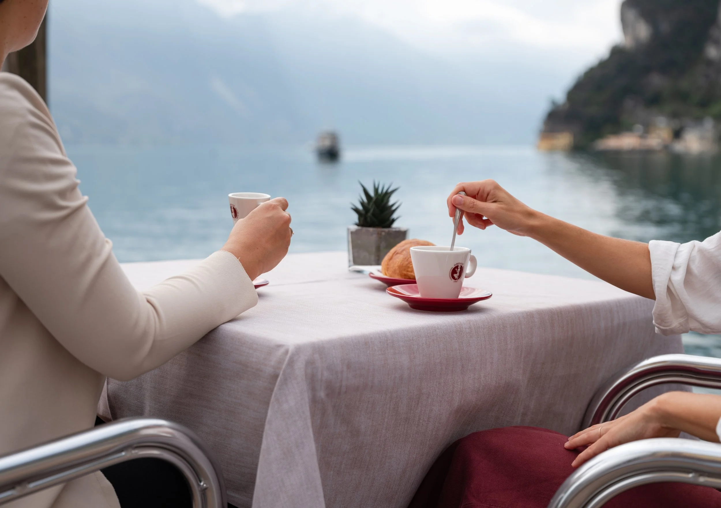 due amiche sedute ad un tavolino di un bar in riva al lago di garda bevono un caffe e un cappuccino omkafe