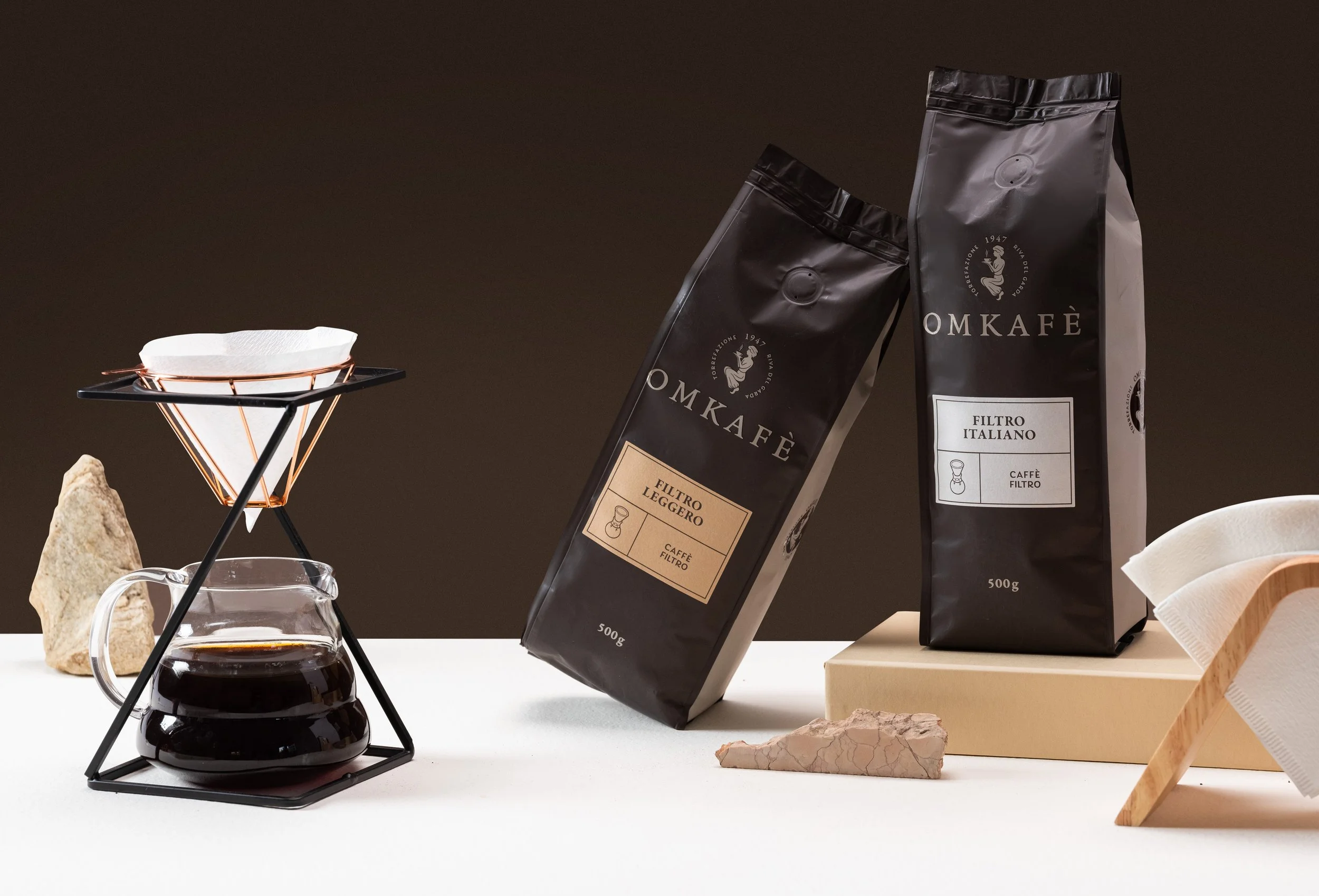 Confezioni di caffè MOMKAFÈ e un set per la preparazione del caffè sul tavolo, con un filtro italiano, una caraffa di vetro con caffè e alcuni pezzi di pietra decorativa.