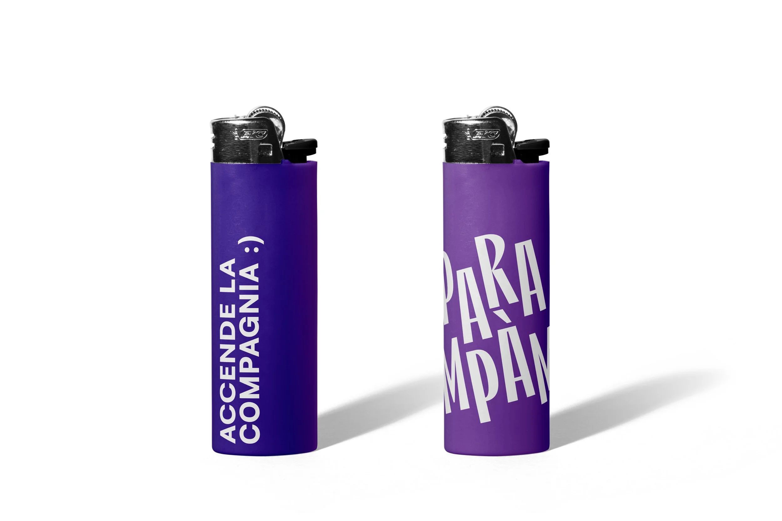 Due accendini di colore viola con scritte in bianco e nero, uno con la scritta 'ACCENDE LA COMPAGNIA' e l'altro con 'PARAMPAMPOLI', entrambi proiettano ombre sullo sfondo bianco.