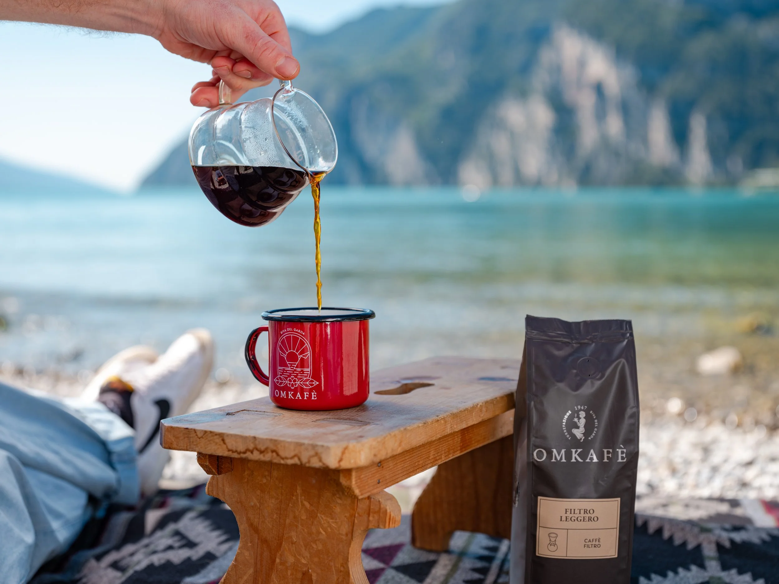 un ragazzo seduto in riva al lago di garda si prepara un caffe in filtro, versa la bevanda nelal tazza di latta e ammira il panorama