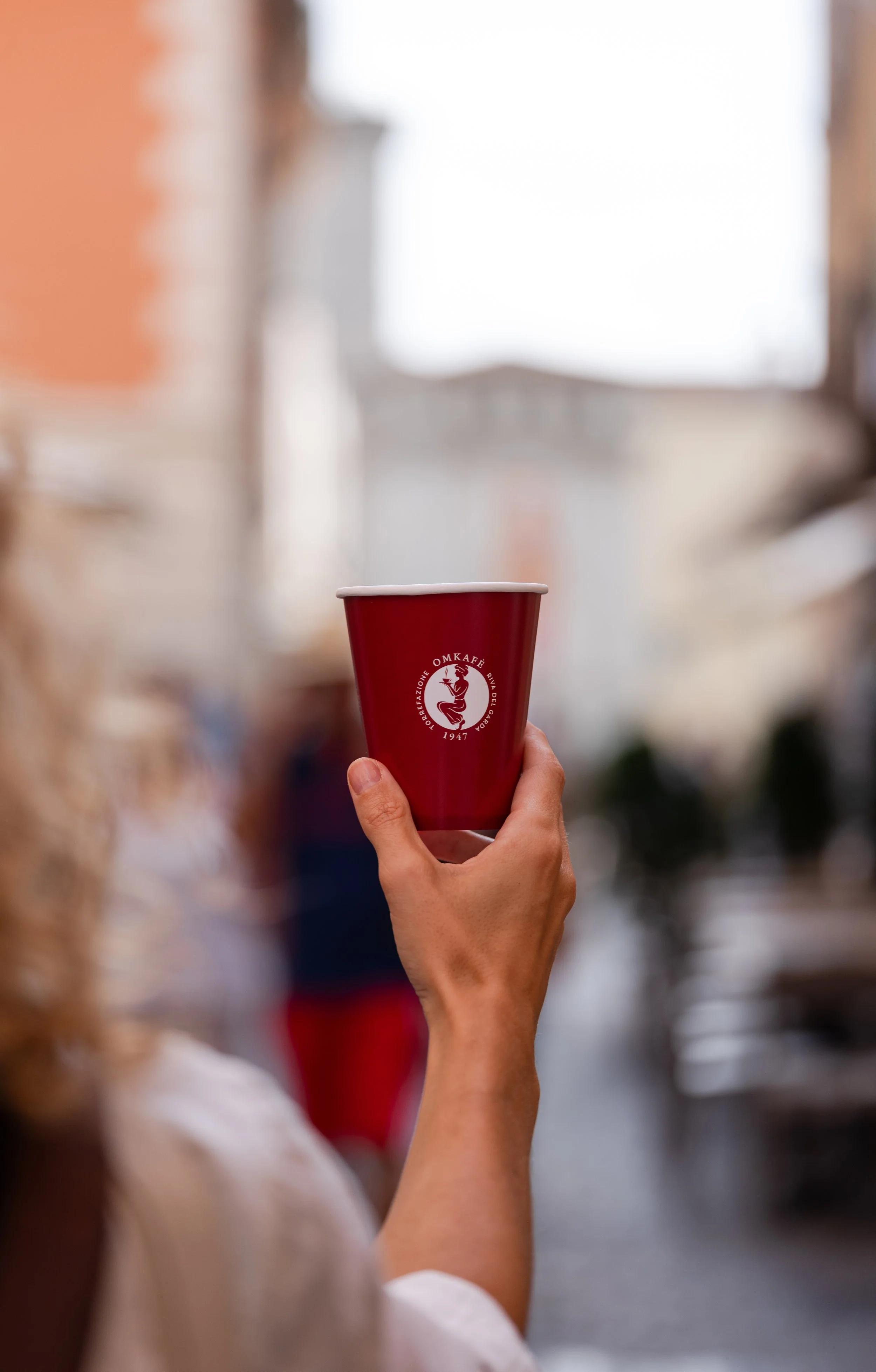 Una mano tiene in piedi una tazza rossa con il logo di un'organizzazione. Sfondo sfocato con altre persone.