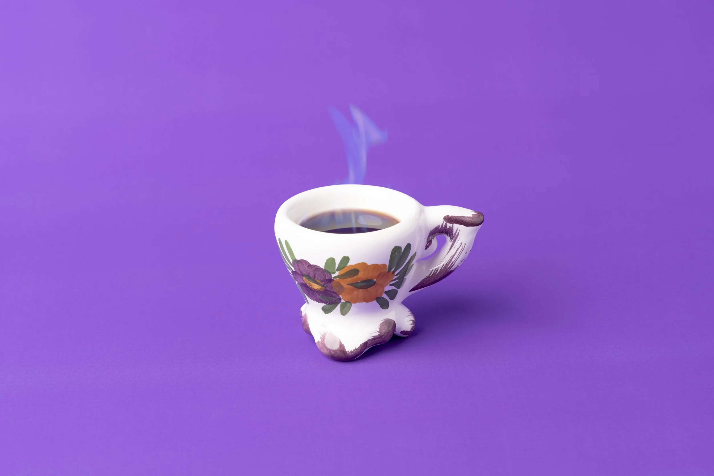 Tazza di porcellana bianca con decorazioni floreali di colore viola, arancione e verde, riempita di caffè nero, posta su uno sfondo viola.