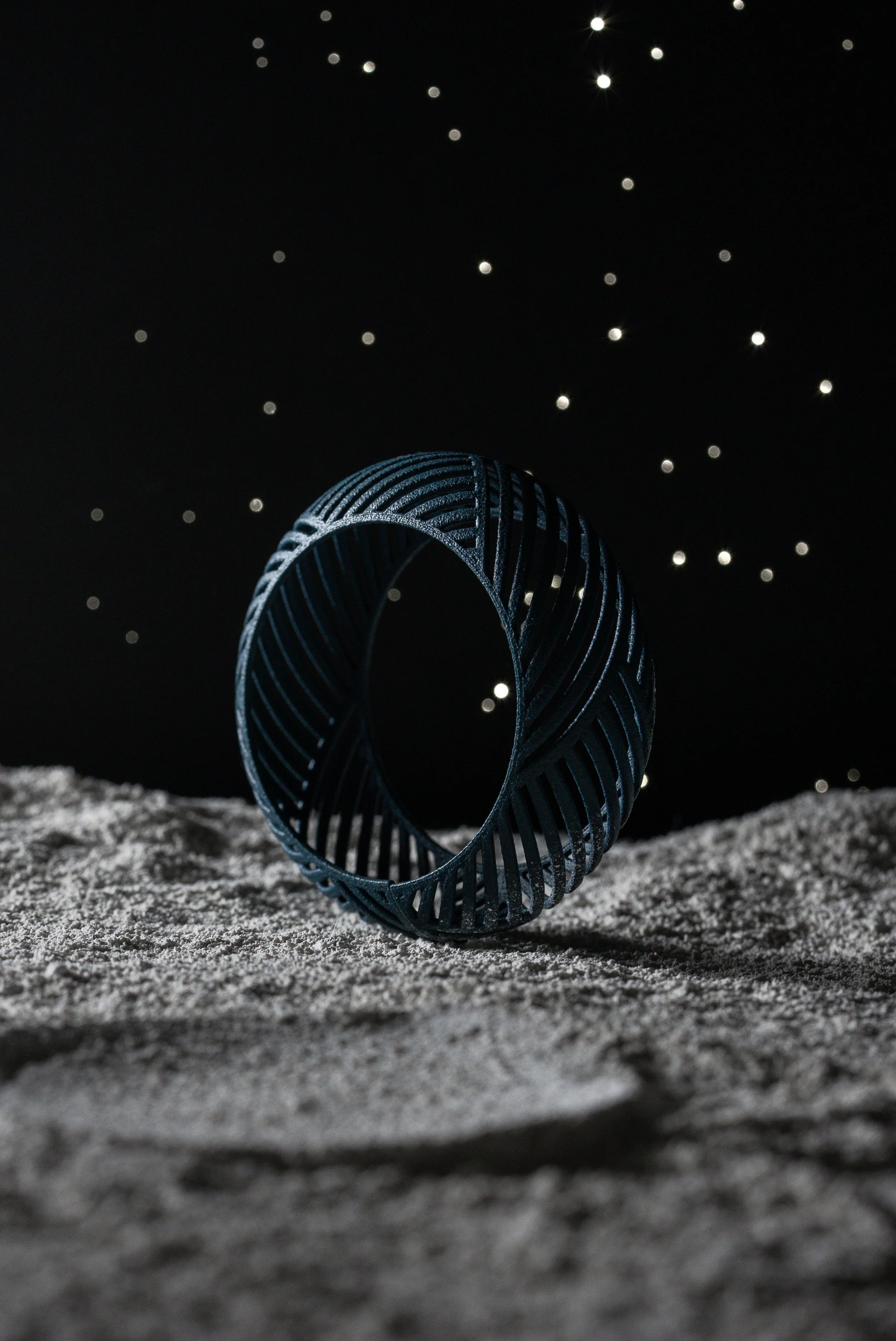 Un anello metallico con design aperto e linee curve, posizionato su una superficie che sembra lette di sabbia, sotto un cielo stellato.