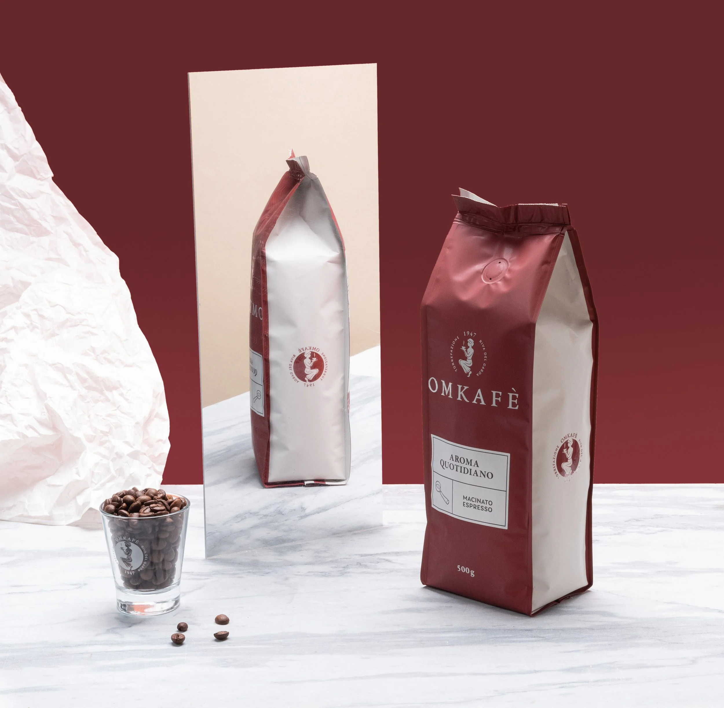 Confezioni di caffè Omkafé e un bicchiere con chicchi di caffè in primo piano su un tavolo di marmo con sfondo rosso.