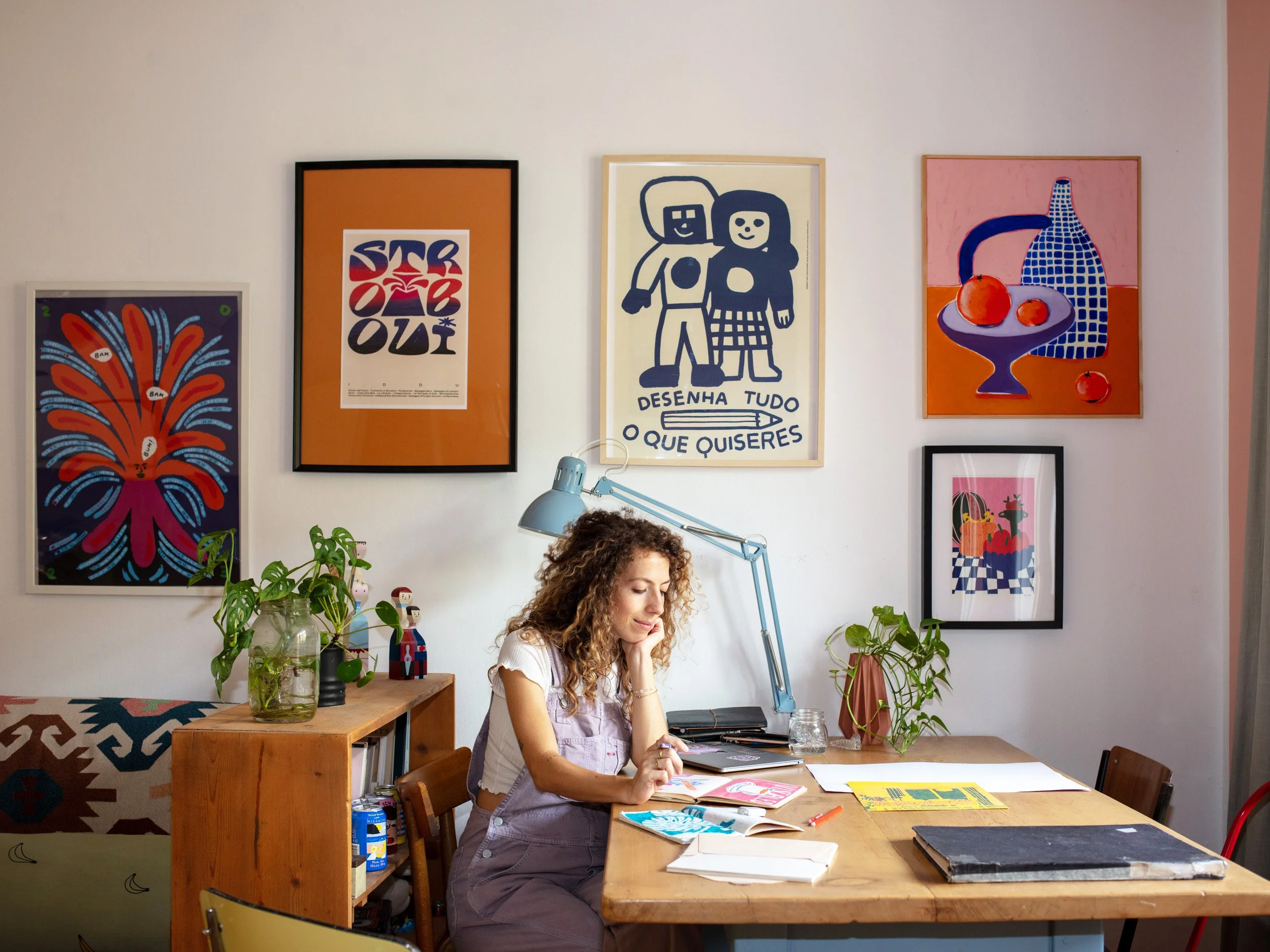 Una donna seduta a un tavolo in un'appartamento decorato con quadri colorati alle pareti e alcune piante, mentre scrive o disegna con materiali artistici.