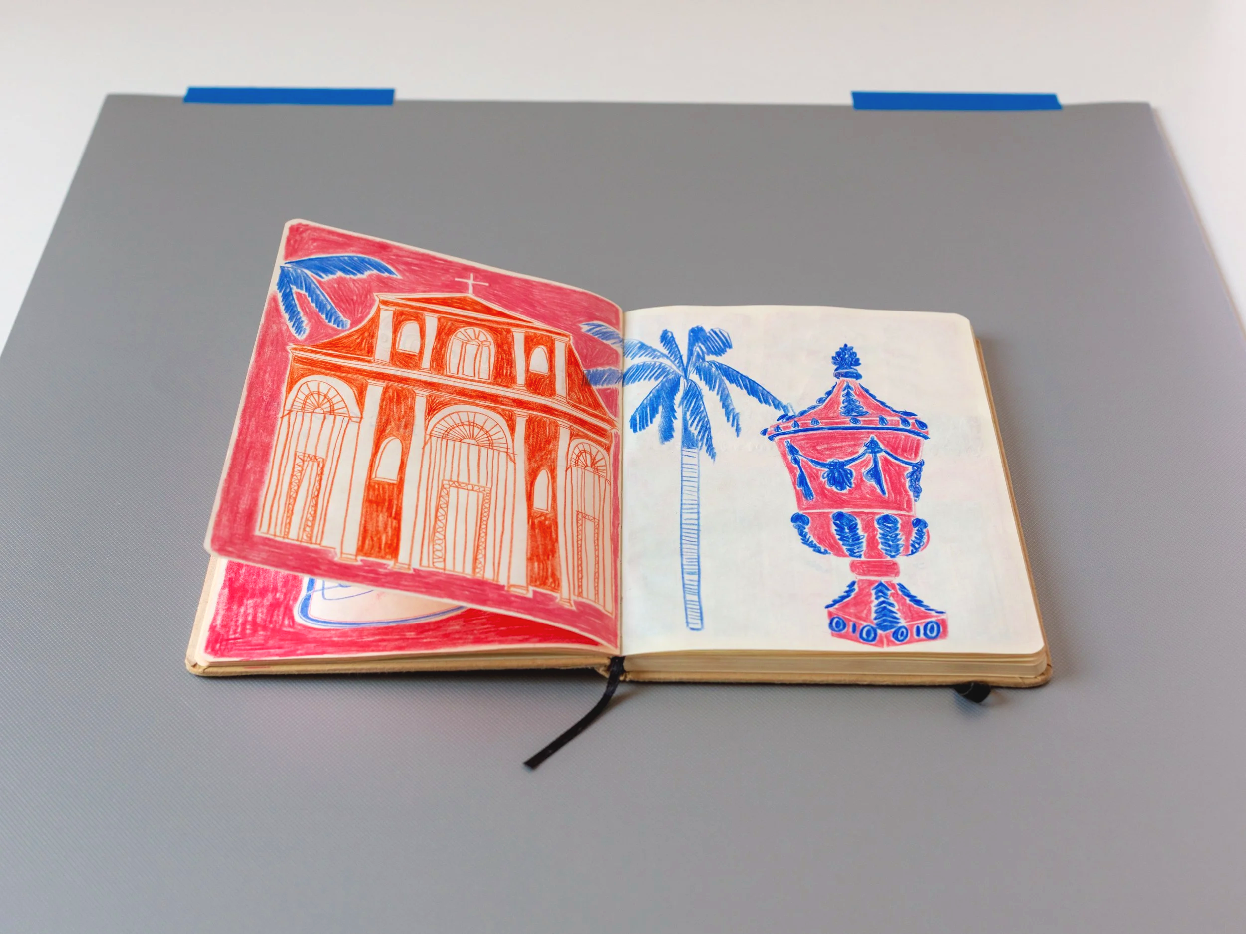 Un taccuino aperto con disegni colorati di edifici e palme blu, presente su una superficie grigia con due pezzi di nastro blu ai lati del piano superiore.