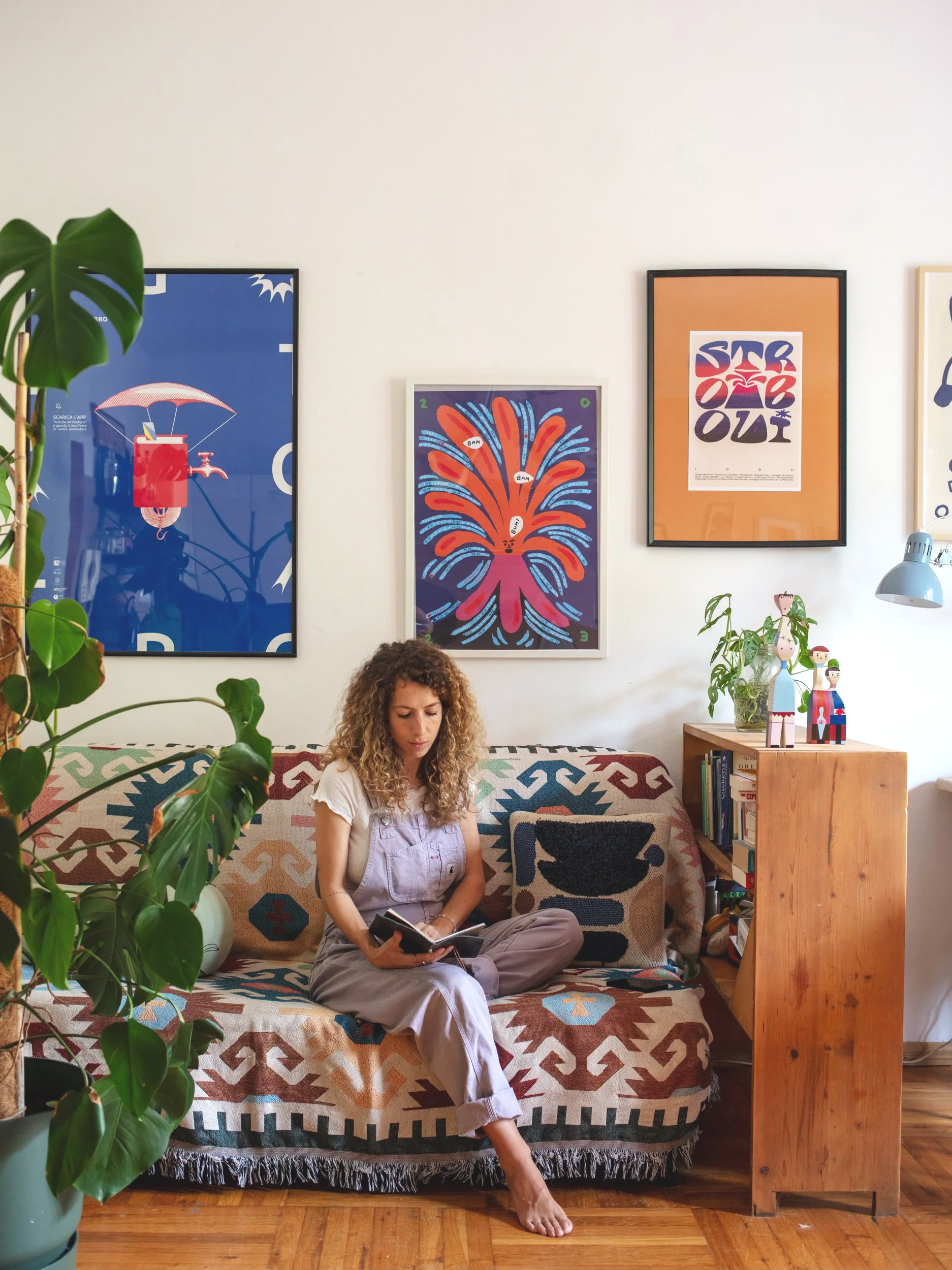 Donna seduta su un divano con motivo geometrico, che legge un libro, in una stanza decorata con poster e piante.