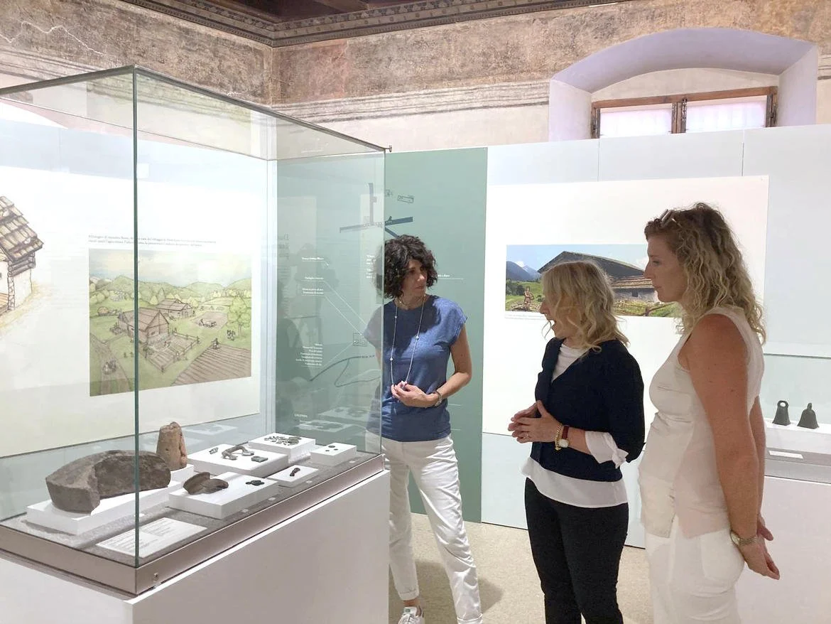 Tre donne in un museo osservano reperti archeologici in una teca di vetro, con disegni di architettura e paesaggi storici sullo sfondo.