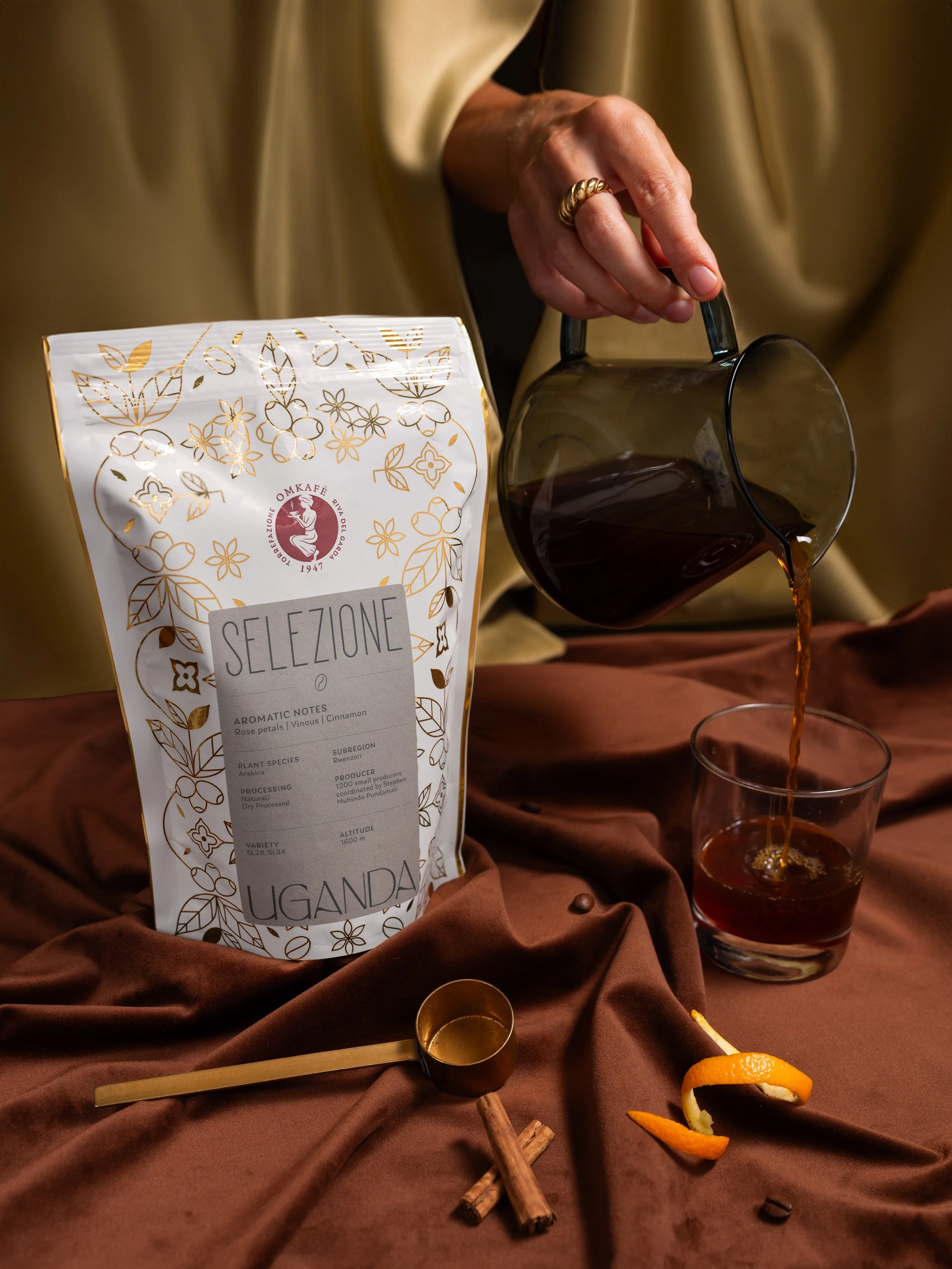 Tazza di tè o caffè scuro mentre viene versato da una brocca, con un sacchetto di tè aromatizzato con note di petali di rosa, profumo proveniente da una confezione sul lato. Sul lato ci sono alcuni bastoncini di cannella, una fetta di arancia e qualche chicco di caffè.