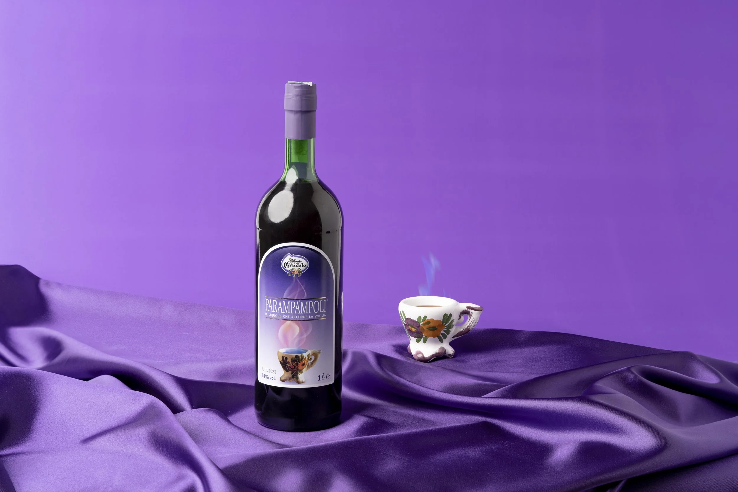 Bottiglia di liquore Paramampoli con una tazza e un piattino decorativi sullo sfondo viola.