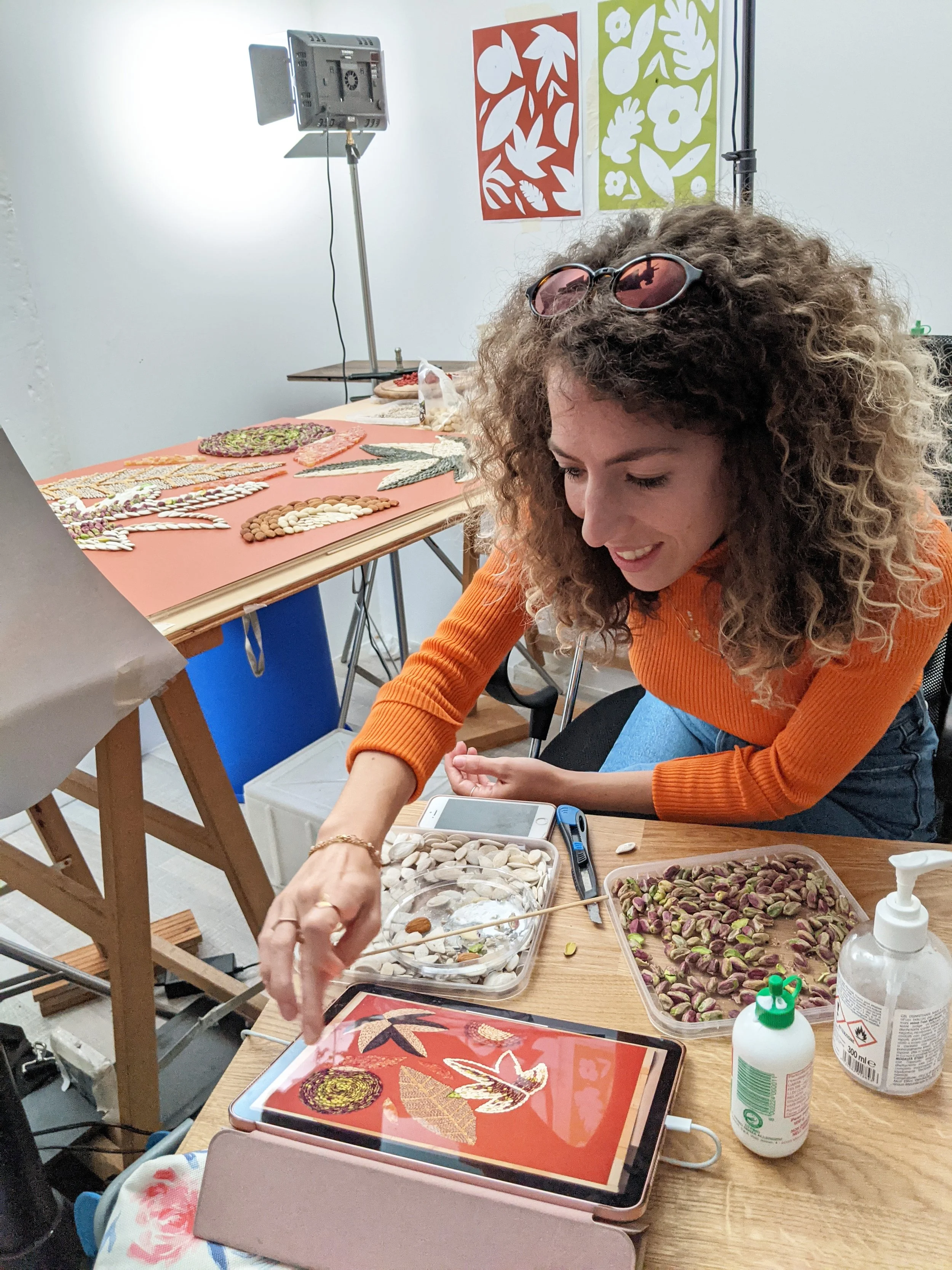 Una donna con capelli ricci scuri indossa occhiali da sole rosa e un maglione arancione. È seduta a un tavolo di lavoro, decorando con semi di pistacchio e altri materiali. Sul tavolo ci sono una tavoletta grafica, barattoli di gel disinfettante e altri strumenti per l'arte. Sullo sfondo, si vedono visivamente due poster con motivi floreali e fogli ricchi di decorazioni di semi.
