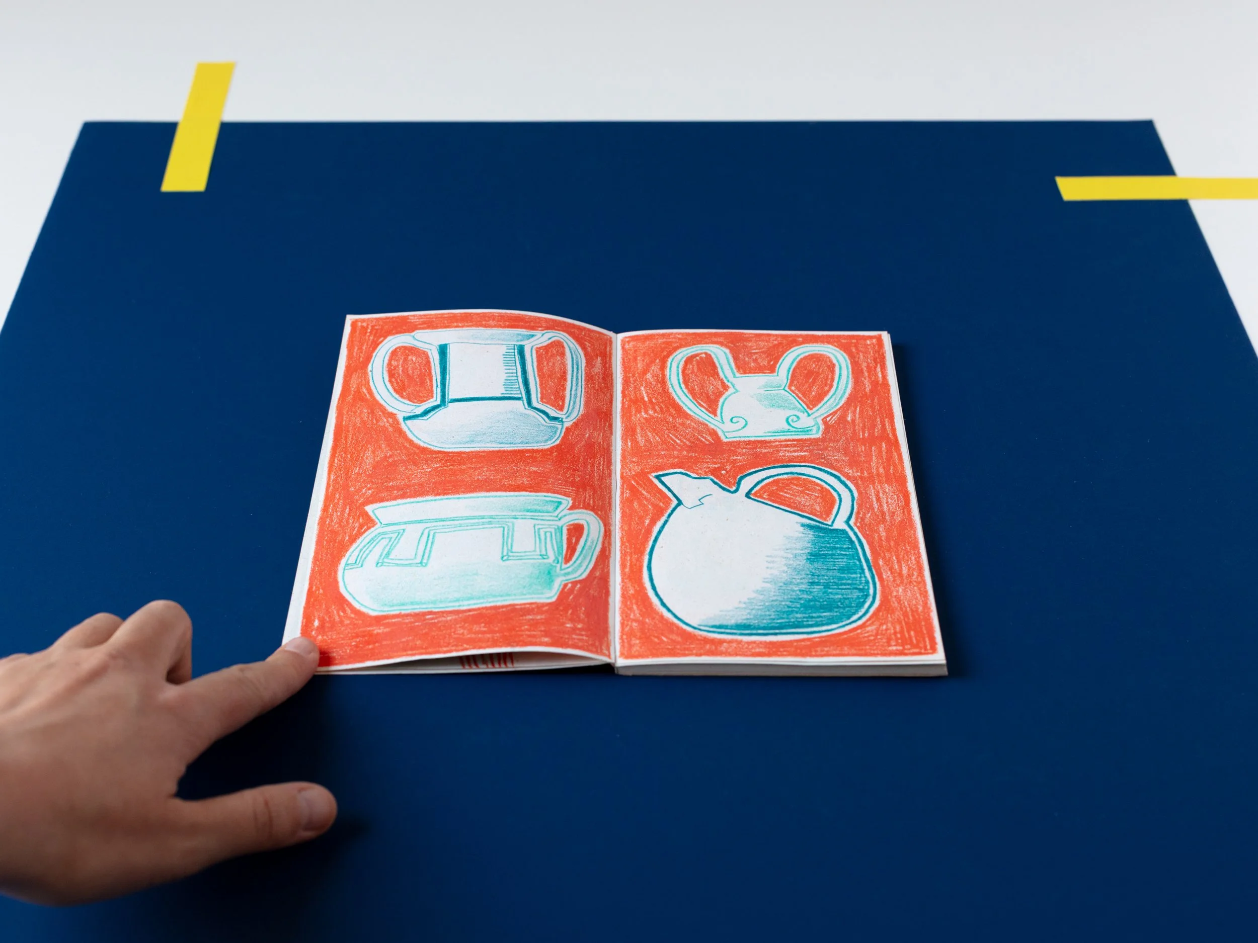 Una mano tiene in mano un foglio illustrato con disegni di vari tipi di teiere, disegnati con colori a pastello, posato su un tavolo blu, con un altro sfondo blu e due strisce di nastro adesivo giallo sulla superficie.