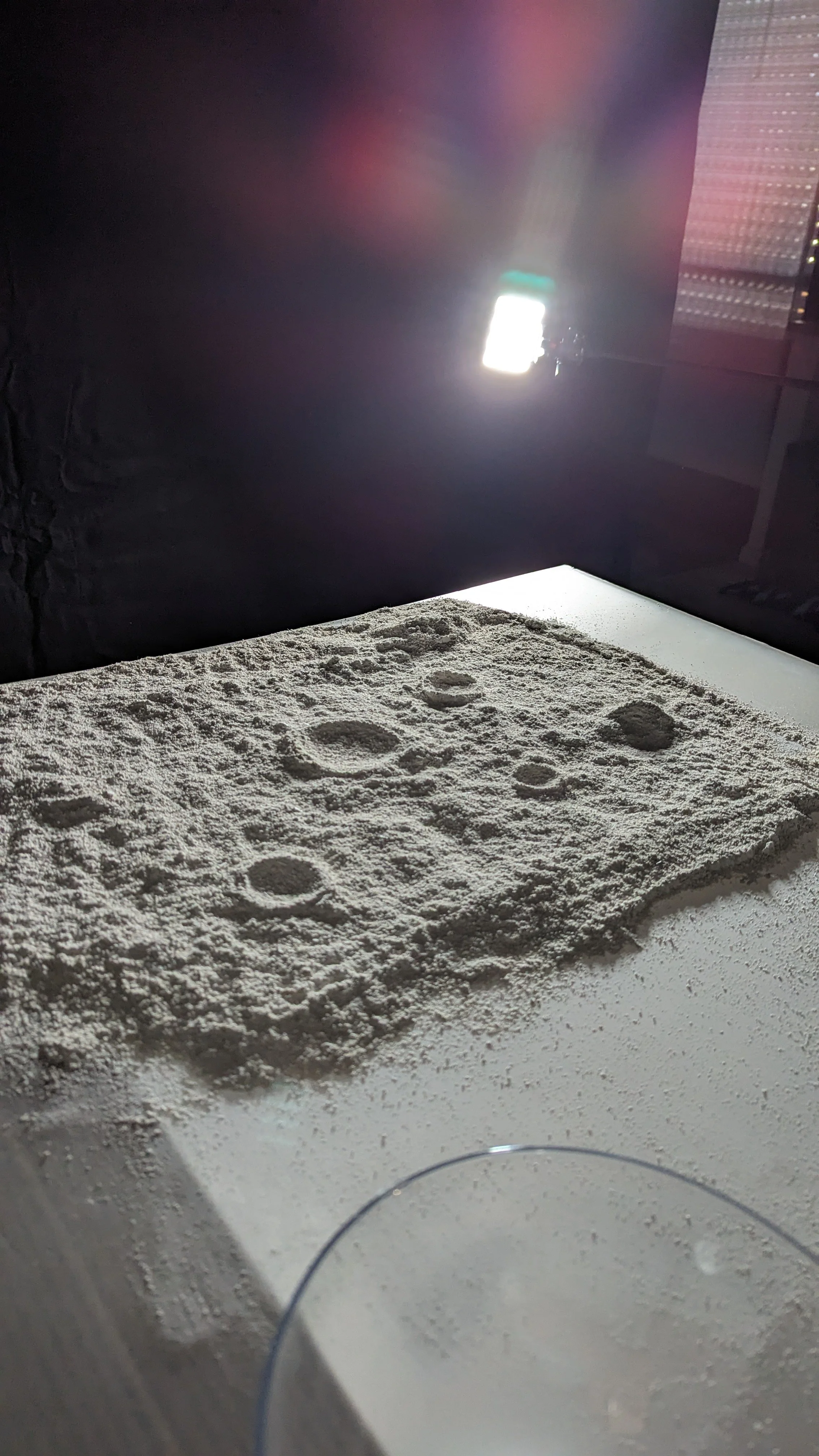 Superficie coperta di farina con alcuni impronte di mani, con sotto un forno a microonde acceso e una lastra di vetro in primo piano.