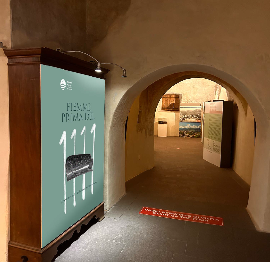 Cartellone al muro con testo in italiano e inglese, che indica l'inizio di un percorso di visita in un museo o esposizione, accompagnato da un'immagine di un mobile antico con trapuntina