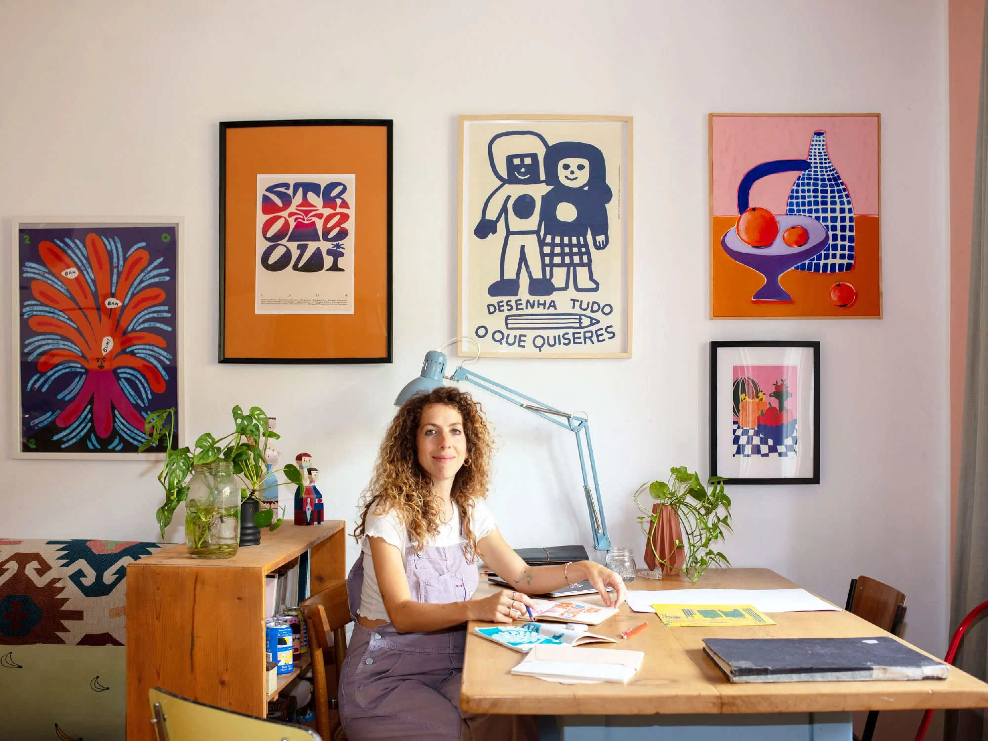 Una donna con capelli ricci seduta a un tavolo con libri e fogli, in un ufficio decorato con quadri colorati e piante, mentre sorride alla fotocamera.