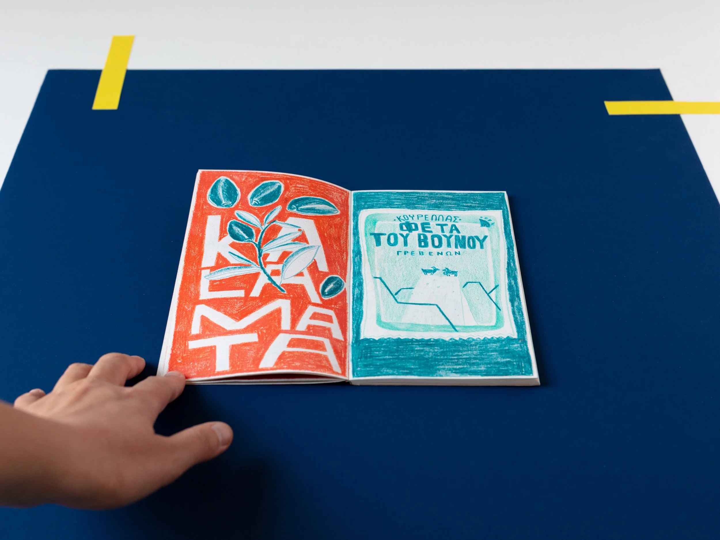 Un libro illustrato aperto posato su un tavolo blu, con una mano che tiene il bordo inferiore. La pagina mostra disegni colorati e testo in greco.
