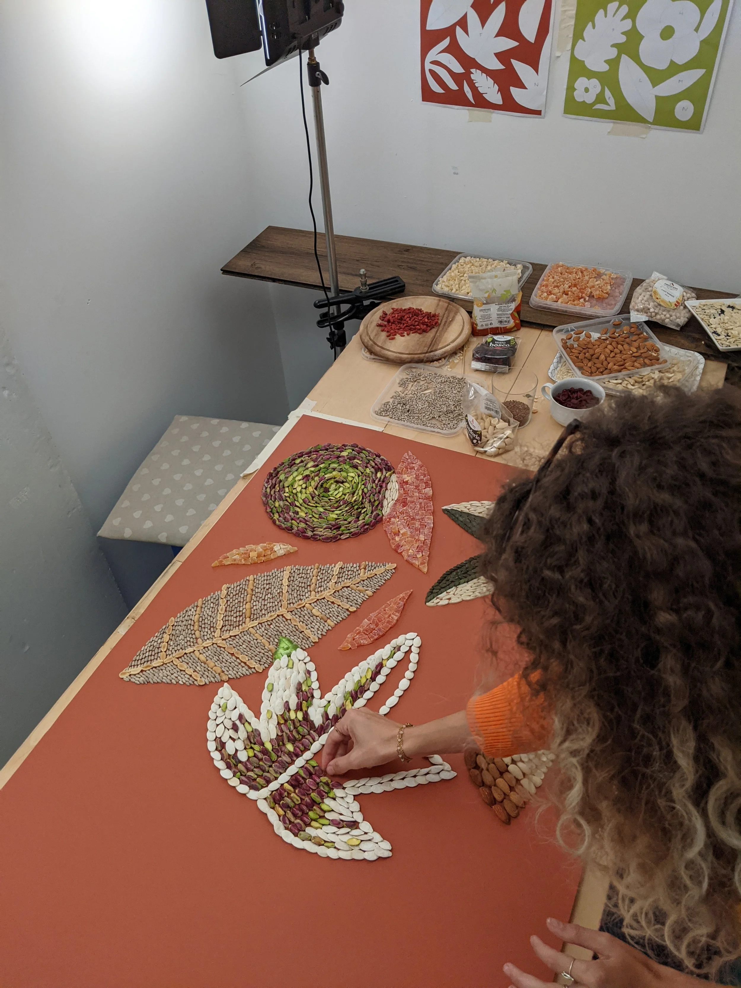 Una donna con capelli ricciato che lavora a un mosaico a forma di farfalla con pezzi di pietre e pasta colorata su un tavolo di lavoro.