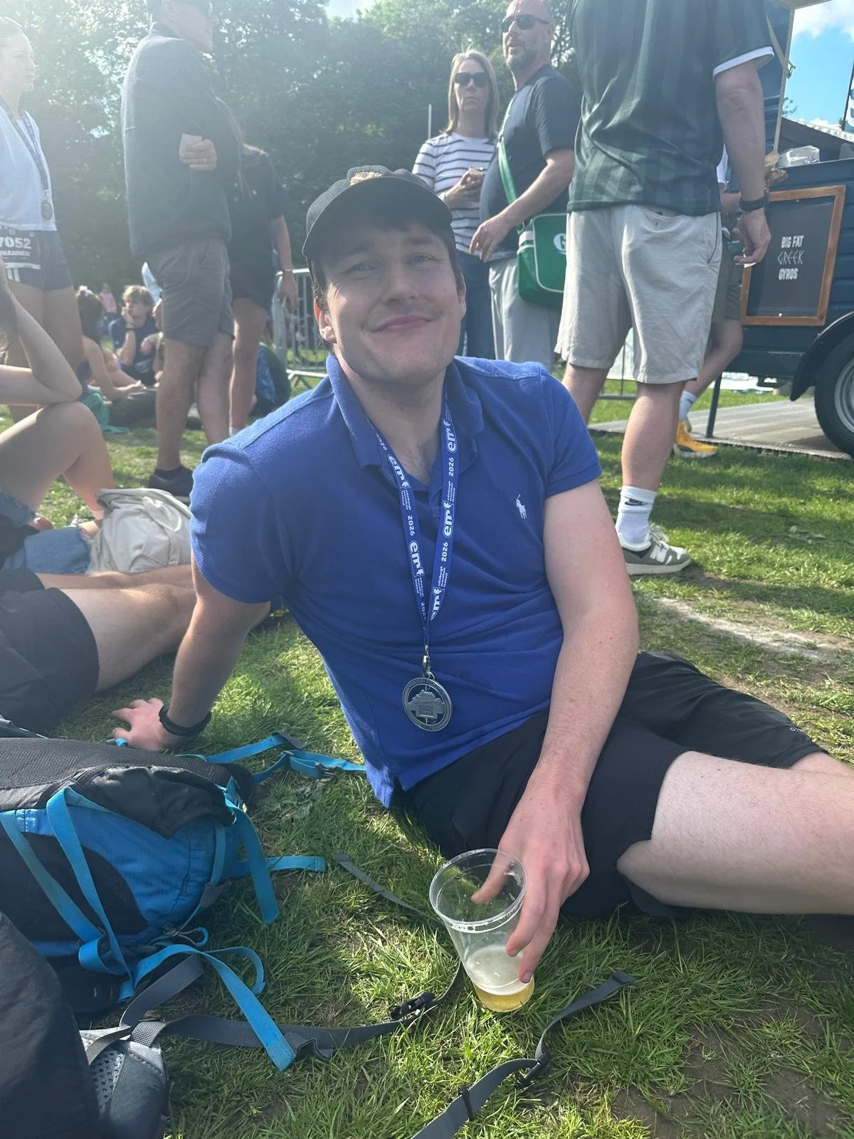 Relaxation Station founder, Jack, after completing the Edinburgh marathon. A man wearing a blue shirt, black shorts, and a black cap is sitting on grass during an outdoor event. He is smiling and holding a clear plastic cup with a beverage.