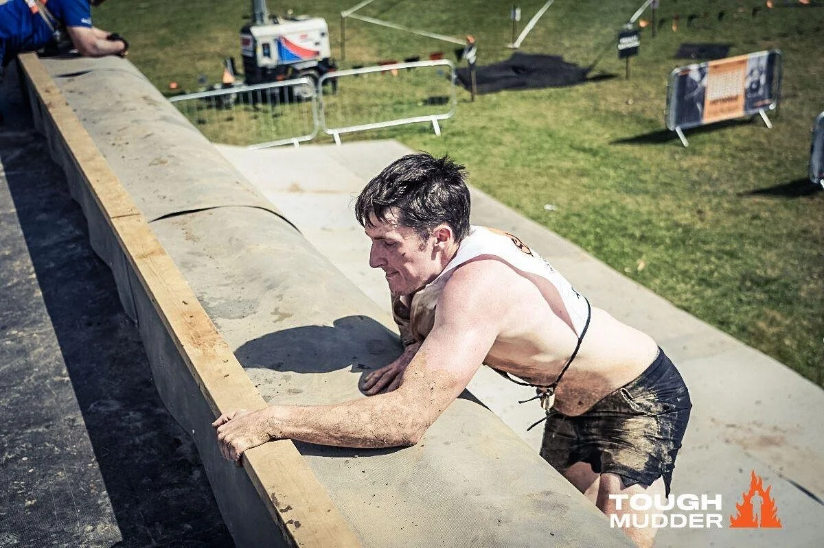 A young man with mud on his arm and shorts is participating in the Tough Mudder obstacle course, climbing over a concrete wall. Our Founder Jack Haygarth participating in the toughest mudder a 24 hour assault course ultra race.