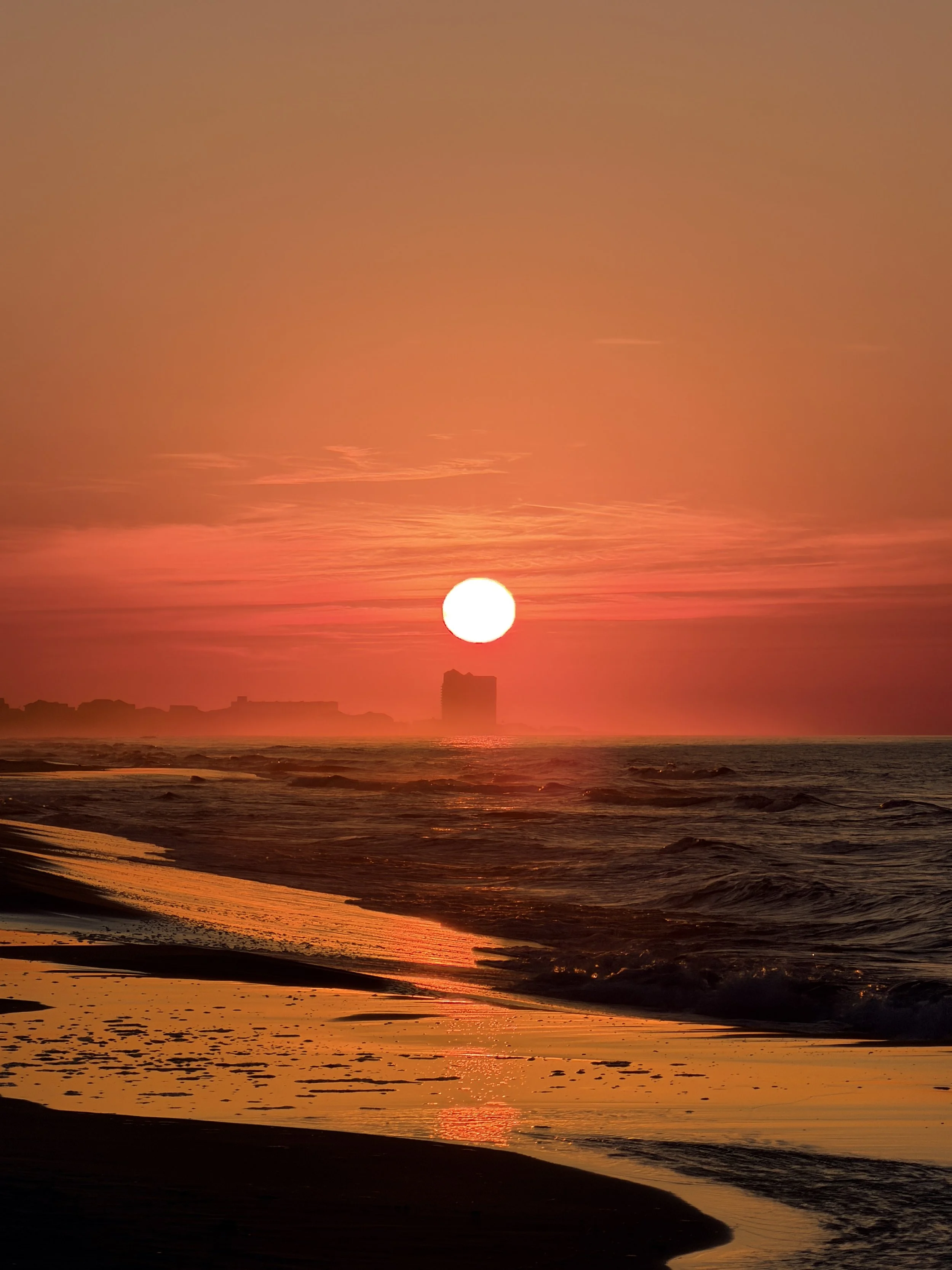 Sunset over the ocean with orange and pink hues, silhouettes of buildings and waves in the foreground.