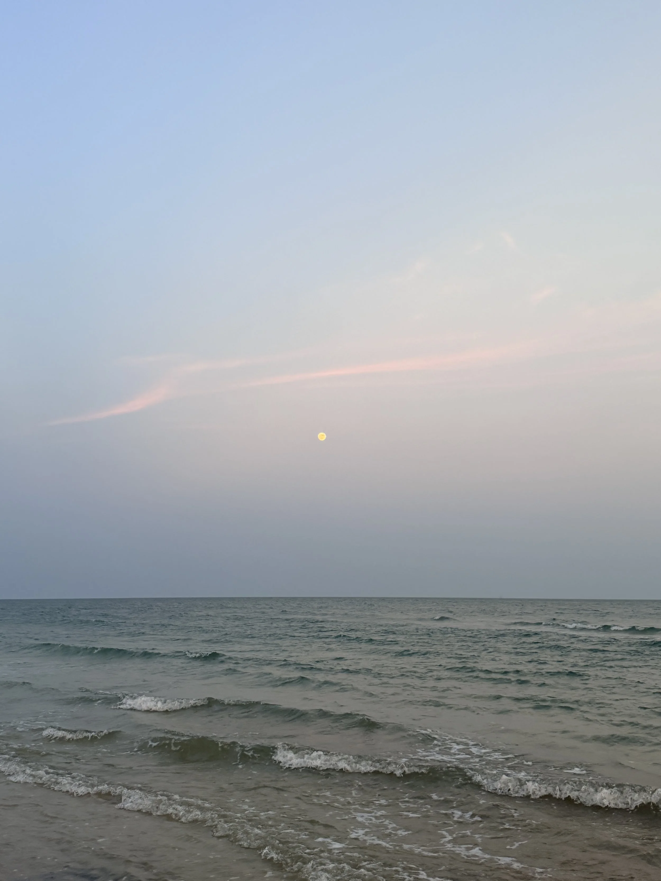 Sea waves on the shore with a setting or rising moon in the sky, showing a calm ocean and a clear sky with some light clouds.