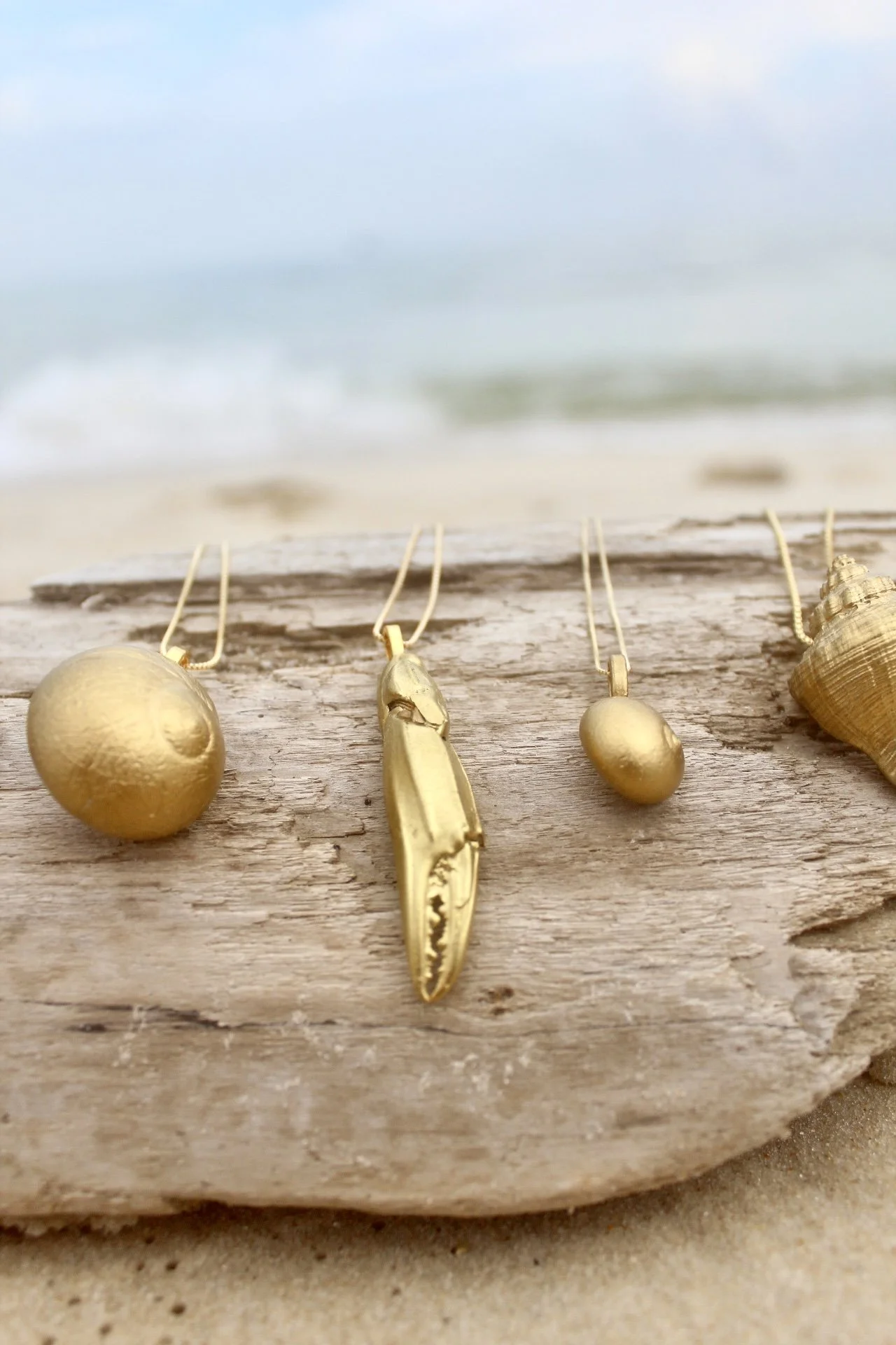 Collection of gold jewelry including earrings, a pendant, and rings displayed on a weathered wooden surface with the beach and ocean in the background.