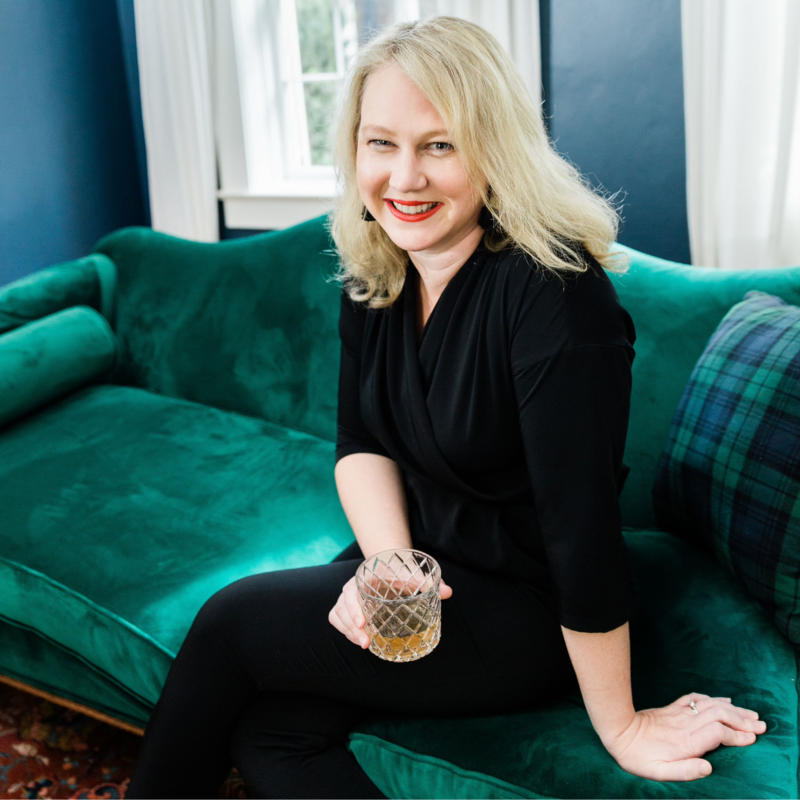 A woman with blonde hair smiling and holding a glass of amber-colored drink, sitting on a green velvet sofa in a well-lit room.