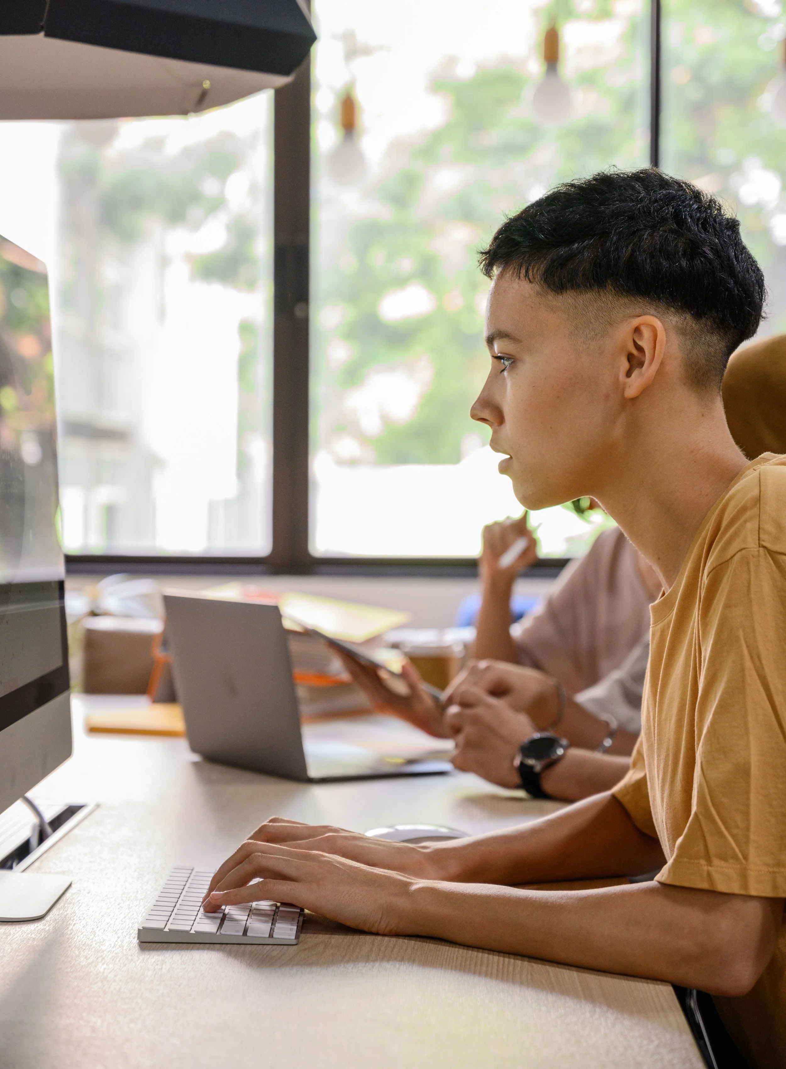 A person with a short haircut working on a computer at a desk in a bright room with large windows.
