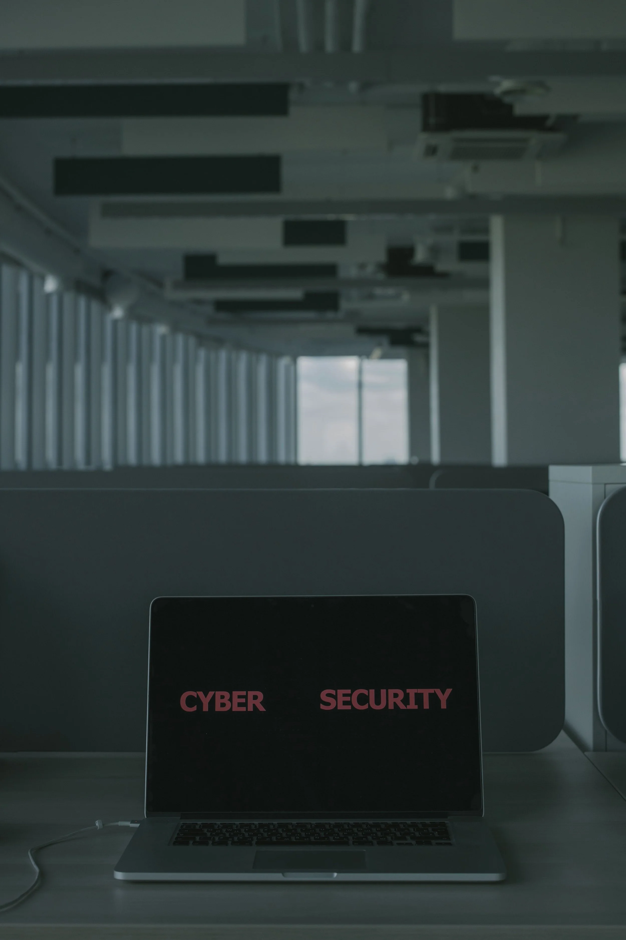 A laptop computer on a desk with the words "CYBER SECURITY" on the screen. The background shows a large window and part of an office environment with ceiling air conditioning units.