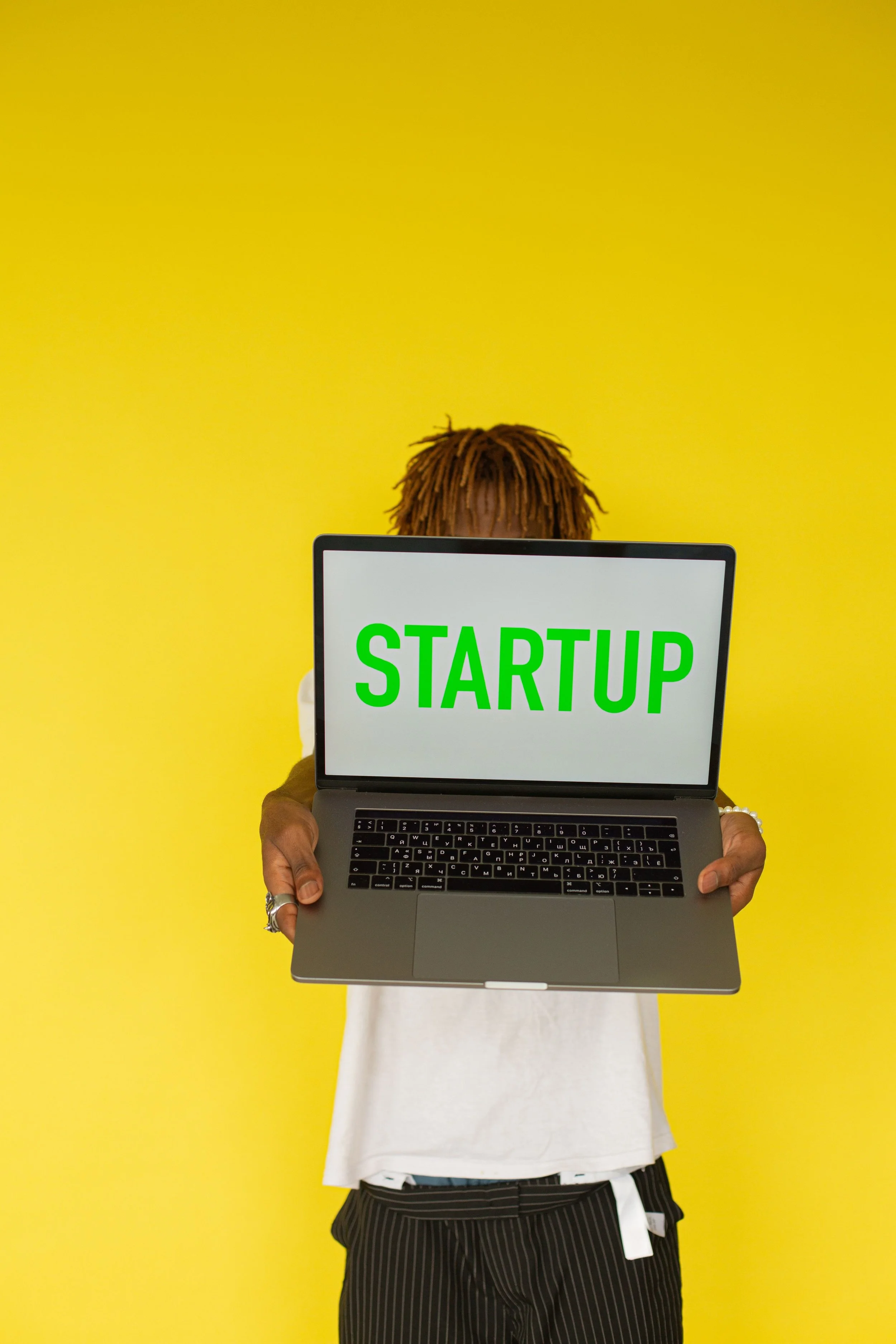 Person with dreadlocks holding a laptop with a sign that says 'STARTUP' in front of their face, standing against a yellow background.