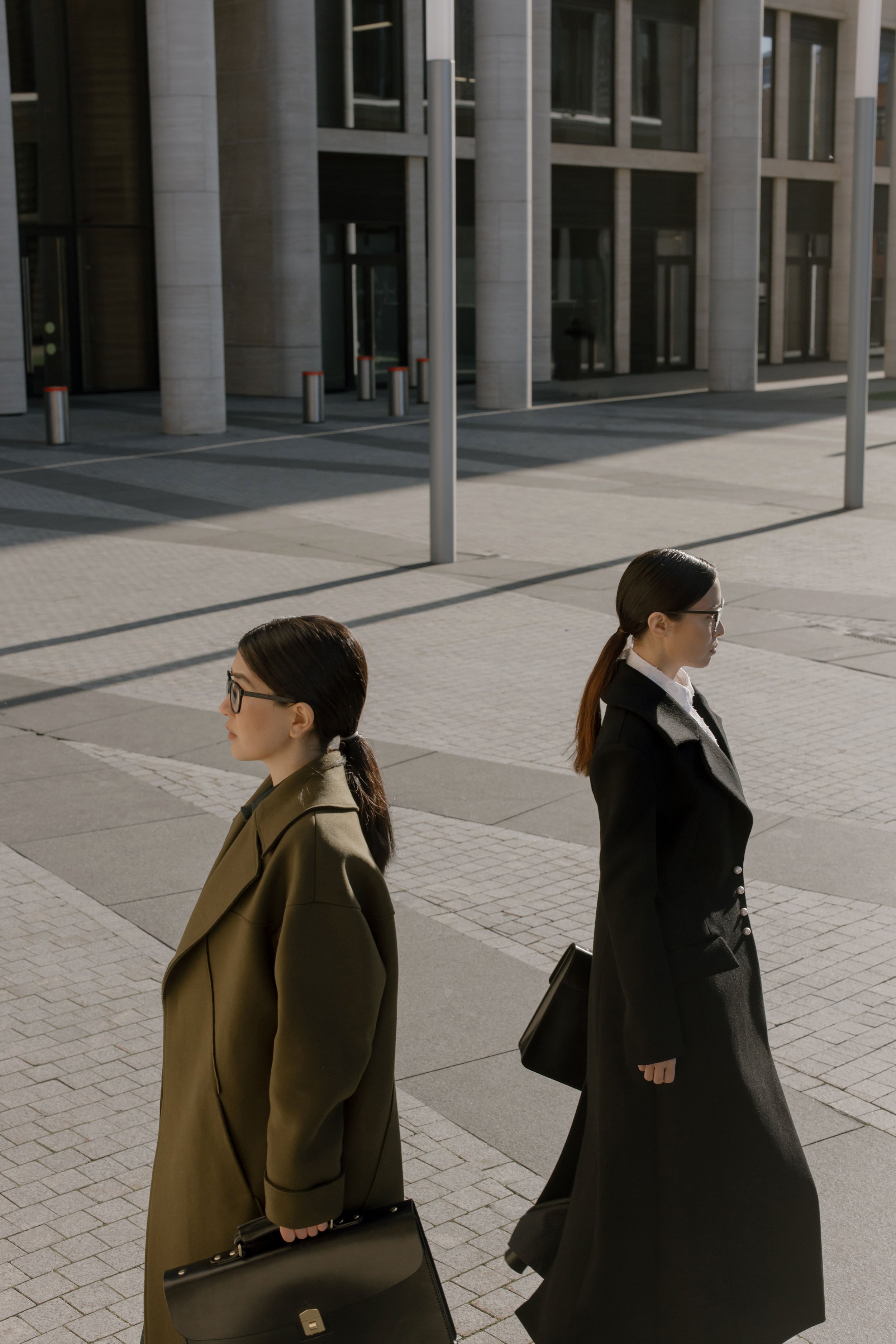 Three women walking outside in business attire, carrying black briefcases, in front of a modern building with large windows and vertical metal poles.