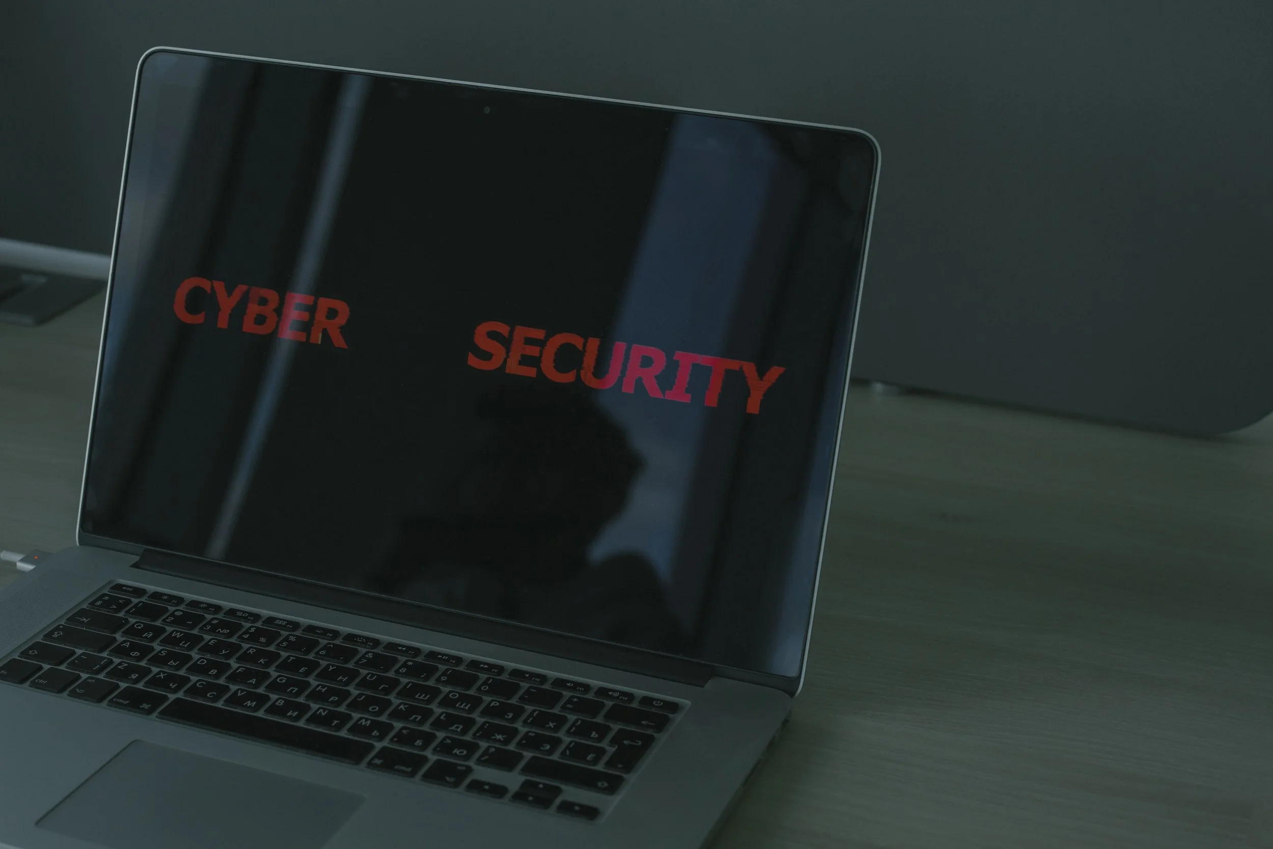A laptop on a desk displaying the words 'CYBER SECURITY' in red and pink text on the screen.