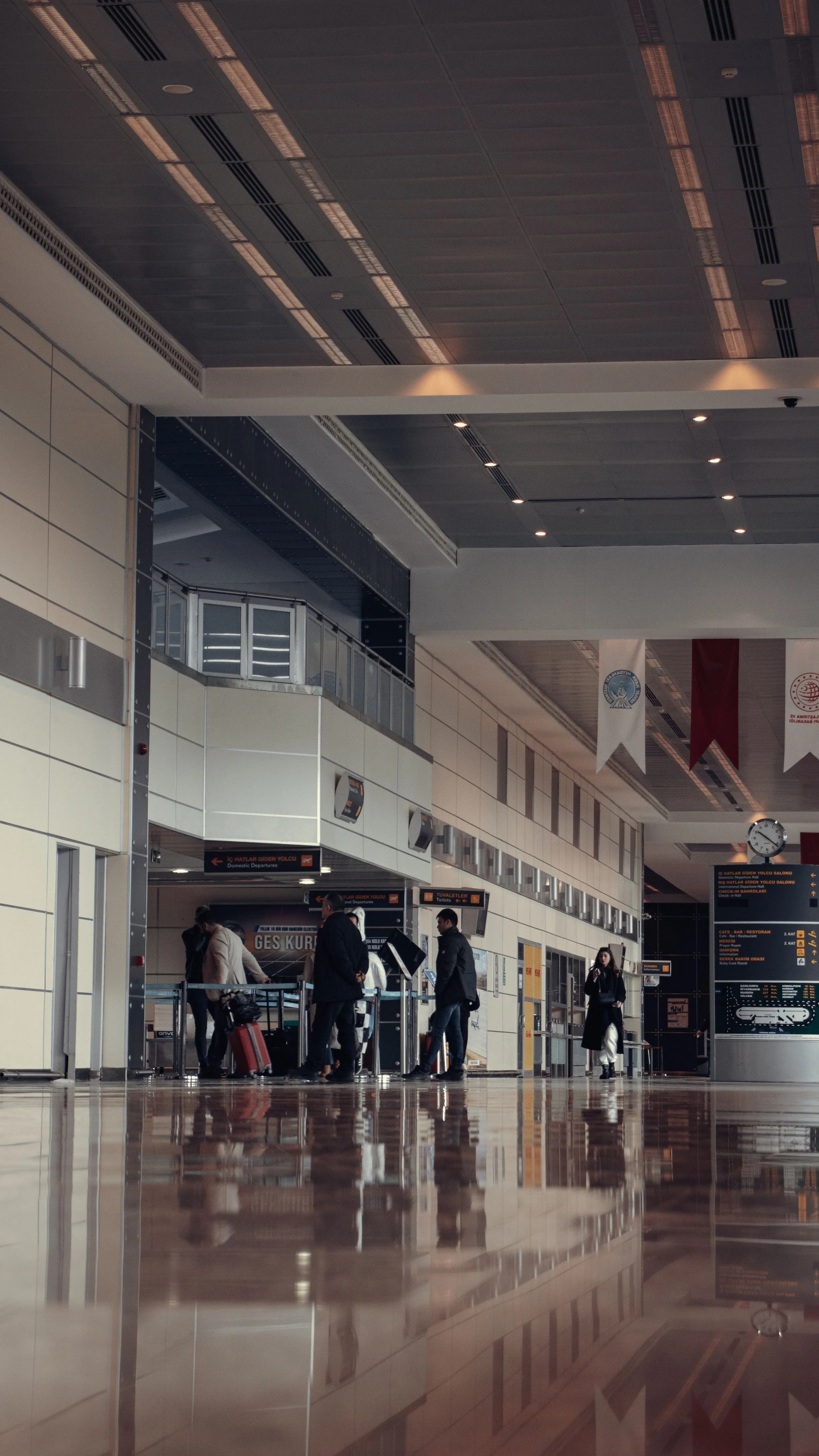 Passengers at an airport check-in counter inside a modern terminal with flags and digital information screens overhead, reflections on polished floor.