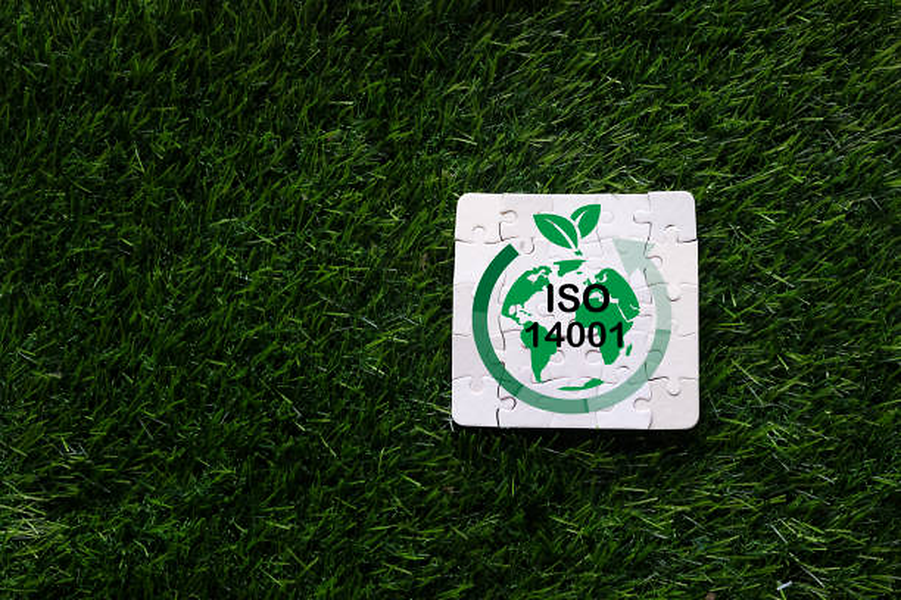 A puzzle piece with a green earth logo, leaves, and the text "ISO 14001" on green grass.