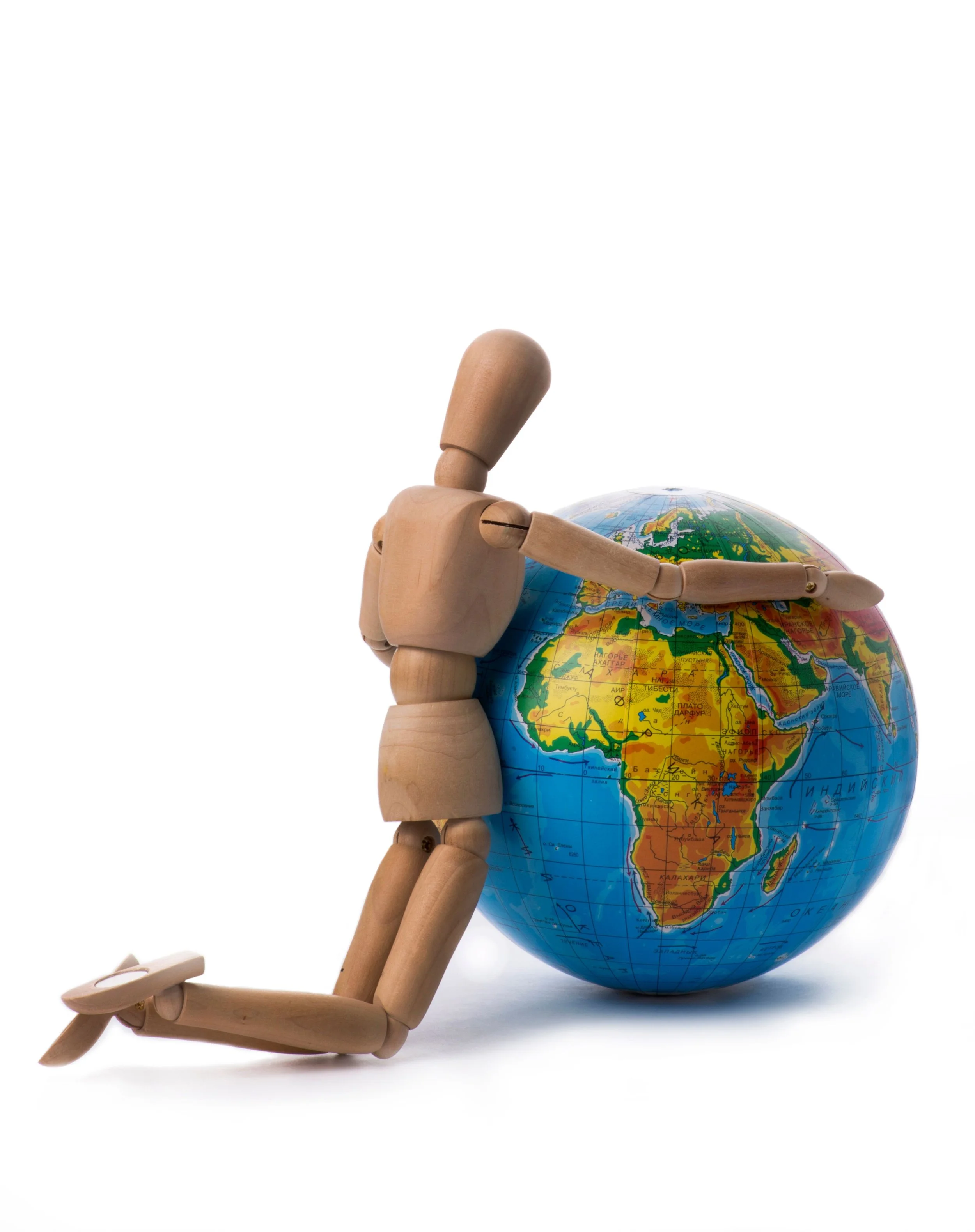 A wooden artist mannequin lying on the floor, with its hand extended and pointing at a globe of the Earth, depicting Africa and surrounding regions, against a white background.